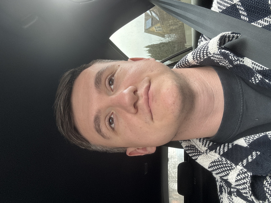 A male individual is observed seated in the passenger side of a vehicle, secured by a black seatbelt across his right shoulder and chest. He possesses short, dark brown hair and is wearing a dark grey crewneck shirt underneath a black and off-white plaid jacket with a dense, woven pattern. His eyes are directed upwards and slightly to the right, and his mouth exhibits a subtle, closed-lip smile.
Through the vehicle's rear passenger window, an exterior environment is visible under overcast conditions. A light-colored, possibly stone or stucco, residential-style building with a distinctive orange-tiled roof is present in the mid-ground. Solar panels are installed on a section of the roof. To the left of the building stands a tall, dark green, coniferous tree. Further to the left, behind the vehicle's interior headrest, indistinct bare tree branches are visible against a grey sky. The immediate ground outside appears dark and unpaved. The vehicle's interior roof lining is dark. This scene is situated in Teufenbach-Katsch, Austria.