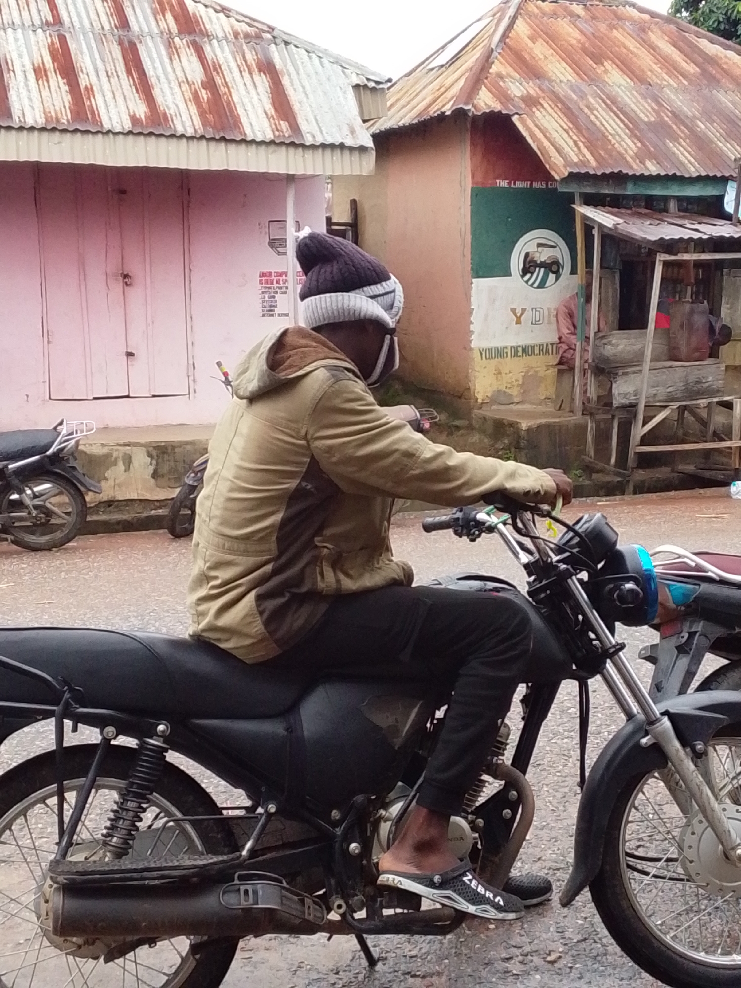 A medium shot captures a Black person sitting on a motorcycle in what appears to be a street scene. The person is wearing a beige jacket with brown accents, black pants, a beanie, and slip-on shoes with "ZEBRA" written on them. They are looking down and holding the motorcycle's handlebars. The motorcycle is black with chrome accents and appears to be parked or stationary. In the background, there are two buildings with corrugated metal roofs. One building is painted pink and has closed doors, while the other is painted in sections of red, green, and white, with political slogans and logos visible. The ground is paved and appears wet. The overall lighting suggests an overcast day.