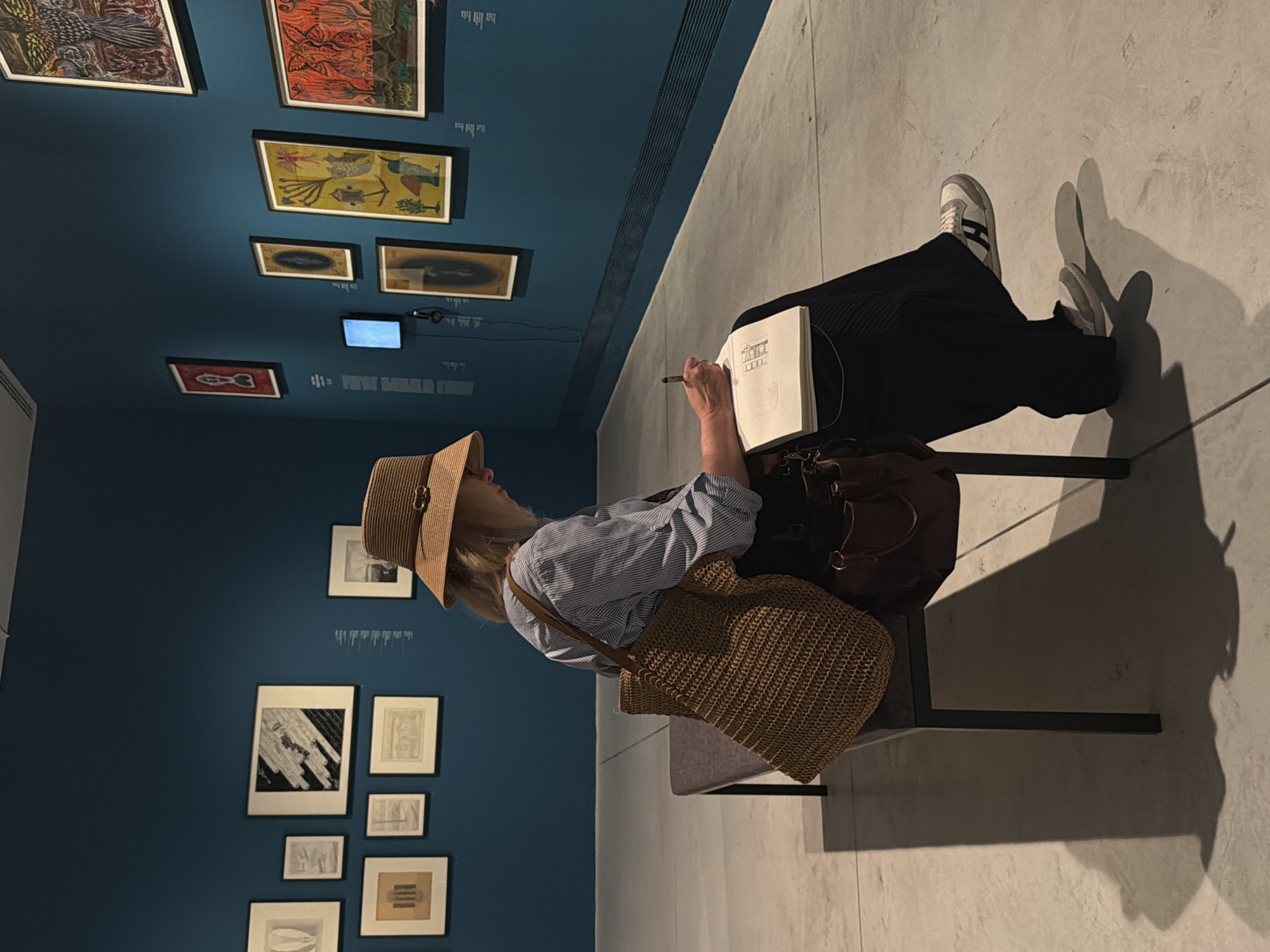 A woman wearing a straw hat and a blue and white striped shirt is sitting on a bench in a museum, drawing in a sketchbook. She is wearing dark pants and athletic shoes. A large straw bag and a brown leather bag are on the bench beside her. The museum walls are painted a deep teal color and are adorned with framed artwork. The floor is a light gray concrete. The image appears to have been taken in the Tel Aviv Museum of Art.