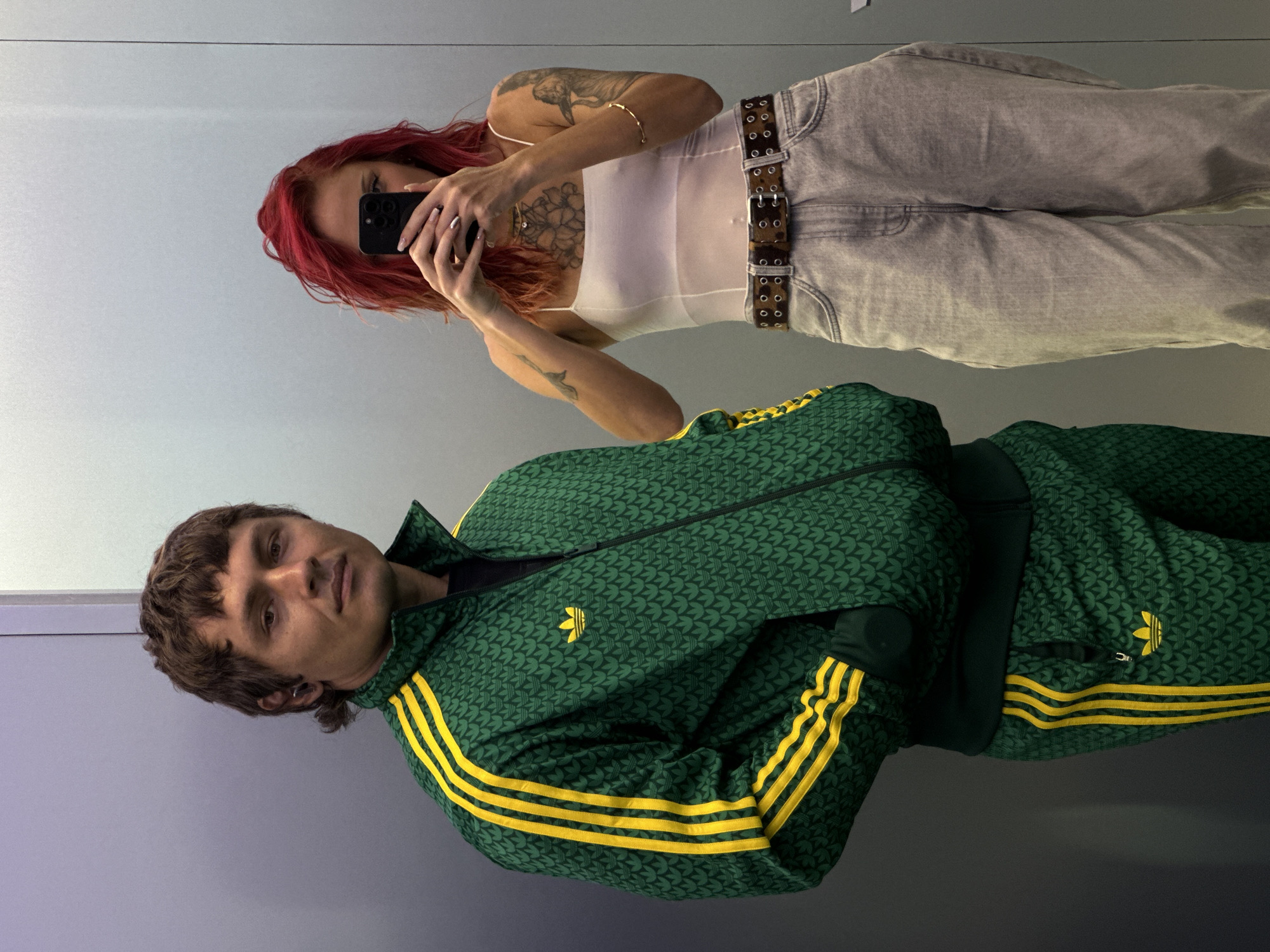 A woman with red hair and tattoos is taking a mirror selfie with her phone. She is wearing a white top, grey pants, and a leopard print belt. Reflected in the mirror, a man wearing a green Adidas tracksuit with yellow stripes is seen. The location of the picture is unknown, with no definitive evidence linking it to Kuta, Indonesia.