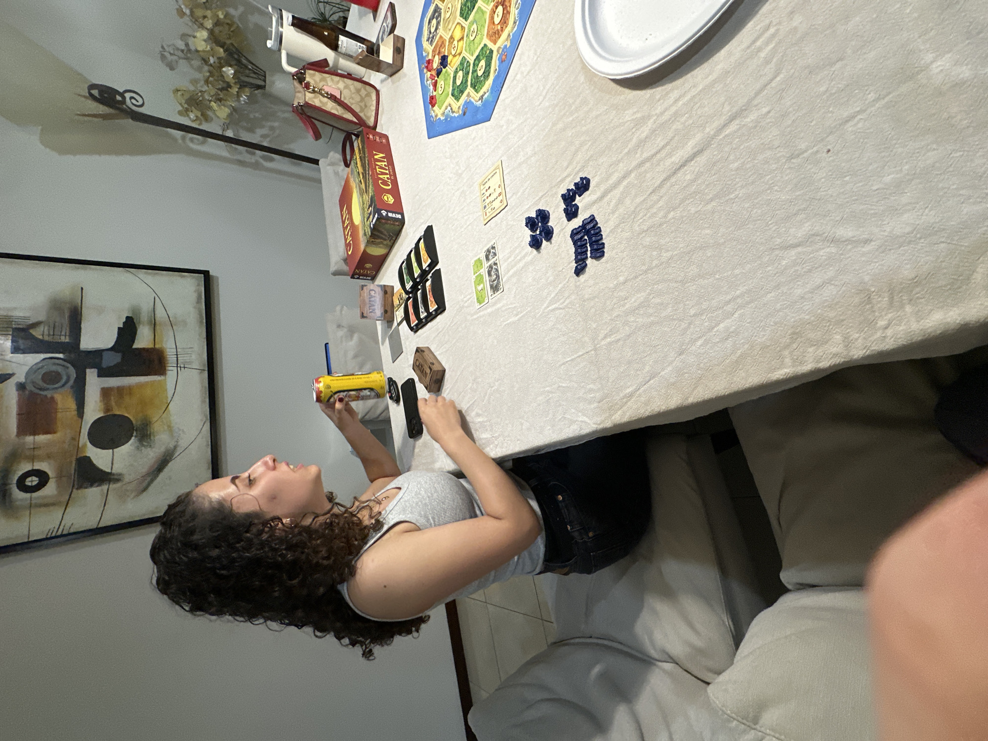 In a room with white walls, a woman with curly dark hair sits at a table covered with a beige tablecloth. She is wearing a light grey tank top and dark jeans, and she is holding a yellow can with a red logo in her right hand. She is looking to her left, speaking. On the table in front of her are several objects, including a "Catan" board game box, game pieces, cards, a phone, and a can of beer. A purse with a red strap and a white pitcher are also visible on the table. The room appears to be a dining area, possibly in San Bernardino Tlaxcalancingo, Mexico, given the context. The overall scene depicts a casual gathering or a game night.