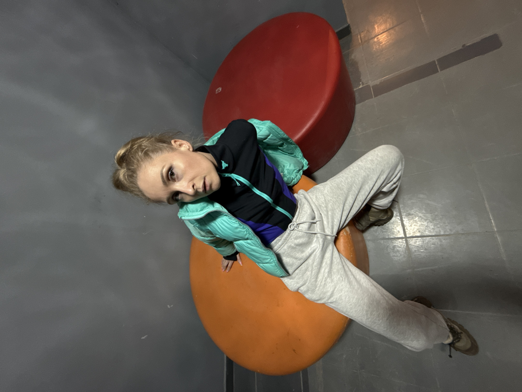 A woman is seen from an overhead angle, seated on an orange, circular stool. She is wearing a light green puffer jacket over a black and blue long-sleeved shirt, with grey sweatpants and dark-colored sneakers. A red, circular stool is positioned behind her. The scene appears to be indoors, possibly in a public area in Chaikovsky, Russia. The floor and walls are grey.