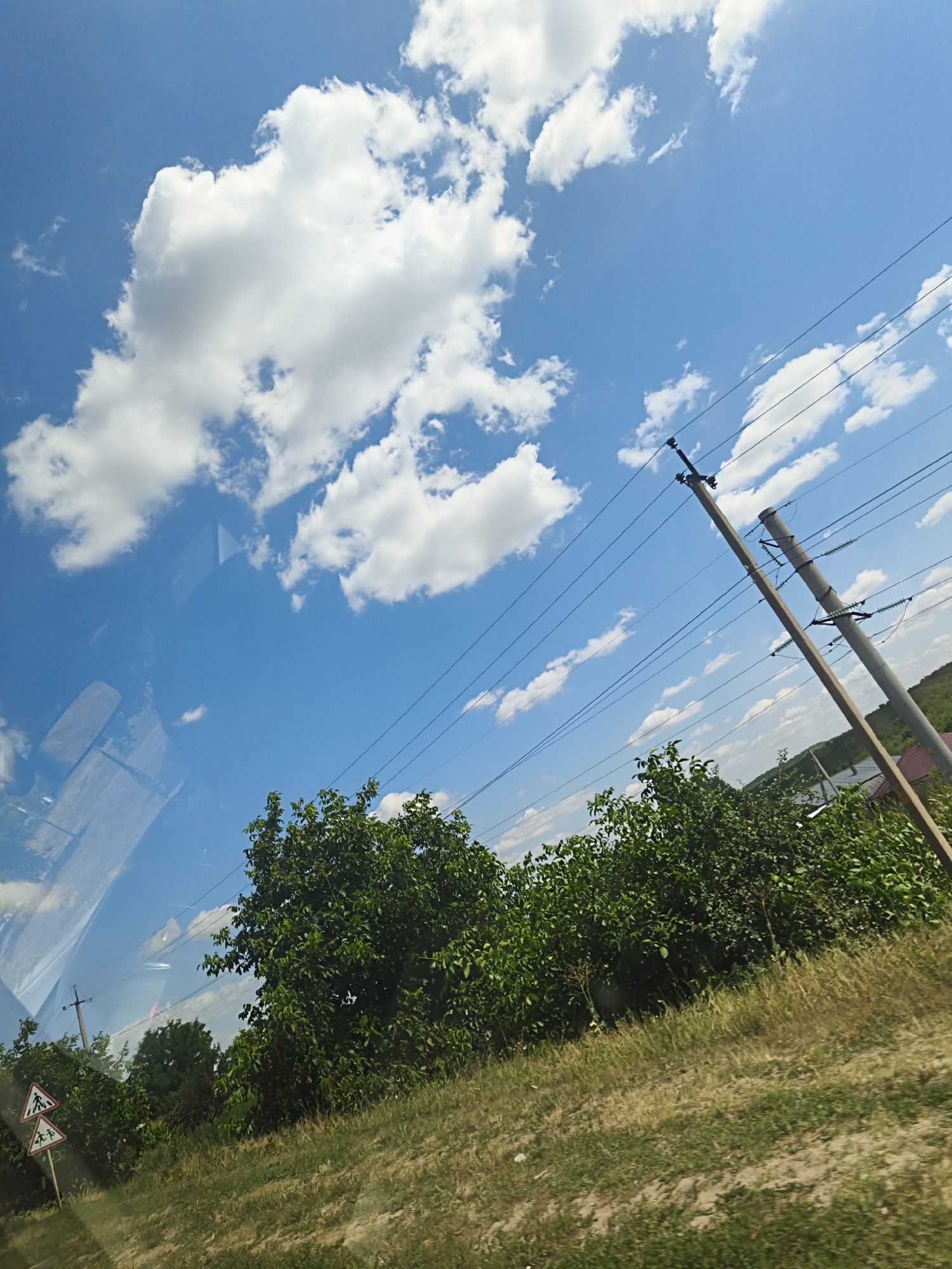 A bright, sunny day is depicted, with a clear blue sky dotted with fluffy white clouds. The scene is viewed from inside a vehicle, as evidenced by reflections on the glass. In the foreground, a grassy verge borders a dirt path. A line of lush green trees and bushes runs along the middle ground. On the right side of the frame, two tall utility poles with electrical wires stretch diagonally across the sky. In the far distance, a few houses and a gently sloping, tree-covered hill can be seen. To the left, two triangular road signs are visible, one depicting a pedestrian crossing. The location is identified as Budești, Moldova.