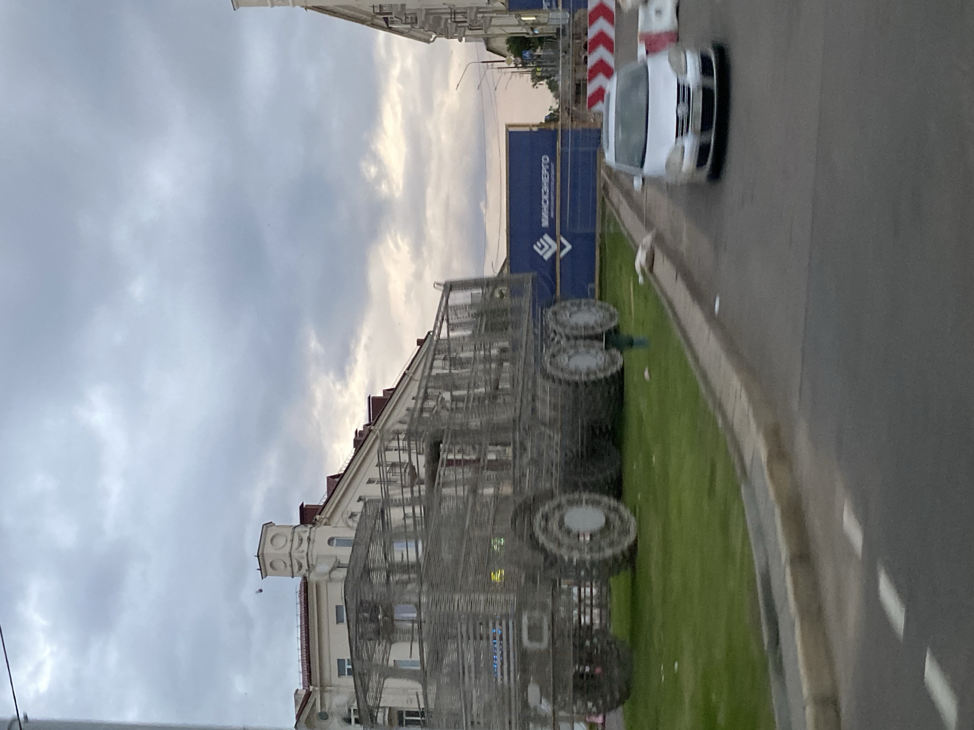This media file shows an outdoor scene that appears to be in a city or town. The primary subjects are a large, wireframe sculpture resembling a bus or tram, a white car, and a building. The sculpture is elaborate and detailed, with what look like large wheels. It is positioned on a grassy area next to a sidewalk and a road. The white car is on the road, moving away from the camera. Behind the sculpture and to the right, there is a blue construction barrier with white text. The building in the background is multi-story with a distinct architectural style, including a tower-like structure on one side. The scene is set under a cloudy sky, suggesting it might be late afternoon or early evening. The overall impression is one of urban infrastructure with a public art installation.
The blue barrier has white text that reads "ВЯЛІКІЯ МЯДВЕДЗІ" which translates to "BIG BEARS" in Belarusian. This is a strong location cue, potentially indicating the name of the installation or a related event.
The angle of the shot is from a slightly elevated perspective, looking down and across the scene. The overall mood is somewhat subdued due to the overcast weather. The presence of a construction barrier suggests ongoing development or repair work in the area.