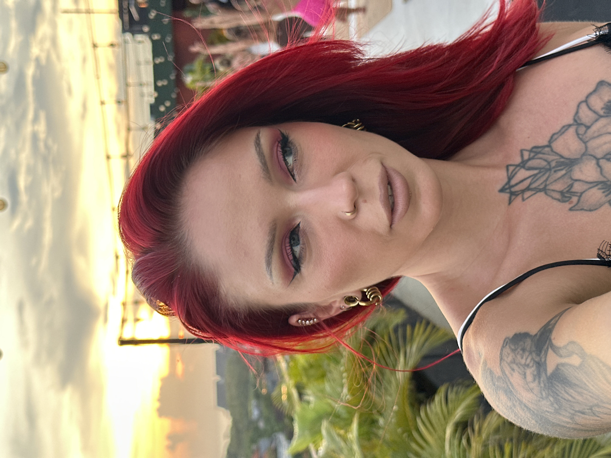 A close-up, candid shot depicts a fair-skinned woman with red hair and visible tattoos on her upper chest and shoulder. She has a nose ring and multiple earrings. Her makeup includes pink eyeshadow and defined eyeliner. The background shows a cityscape during sunset, with palm trees visible in the lower portion. The location is identified as Tibubeneng, Indonesia, based on the visual cues of the tropical setting and urban environment.