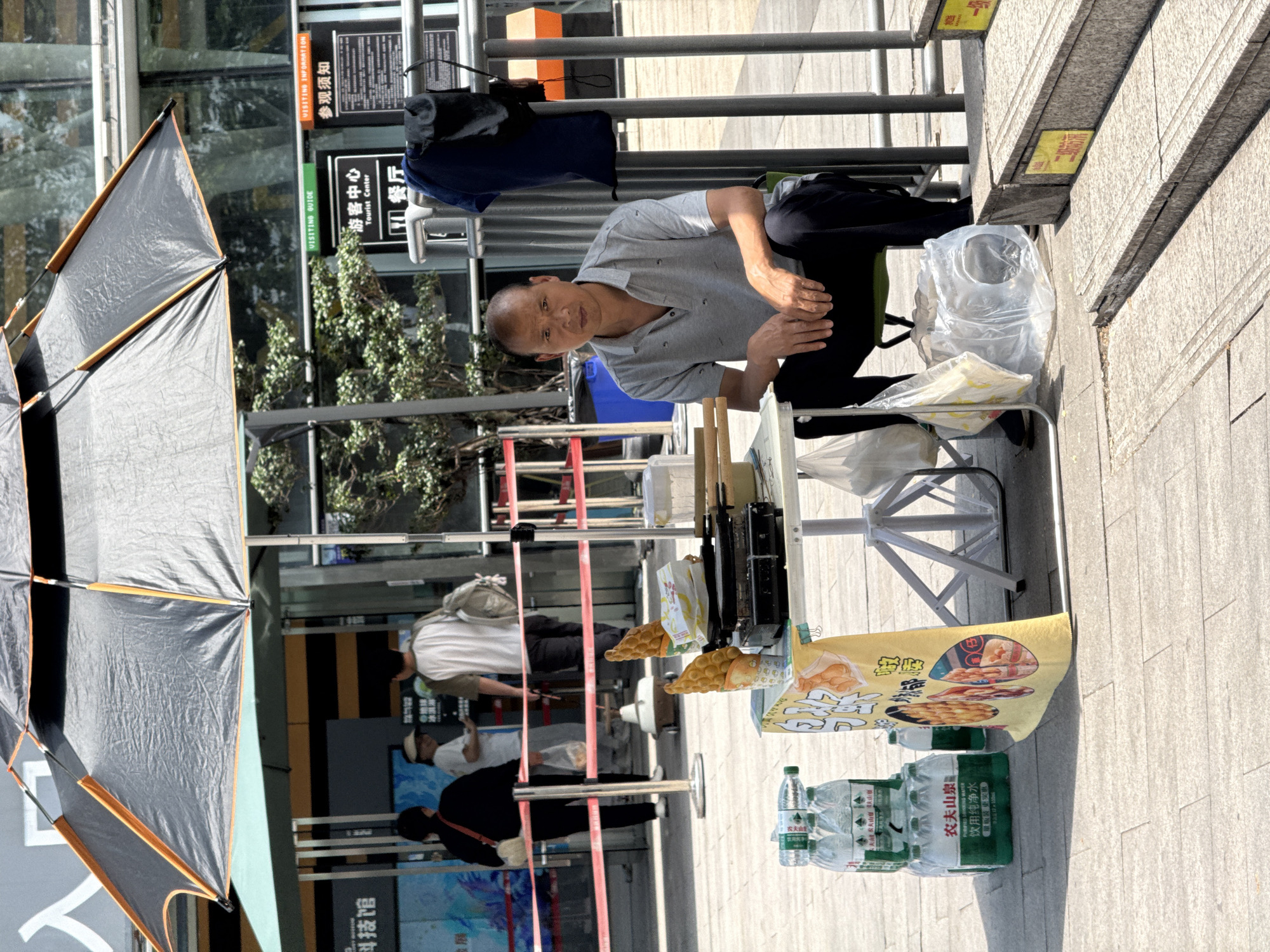 A street vendor operates a stall in 江北城街道, China. An Asian man squats behind the stall, looking towards the camera. The stall has a black waffle iron, cooked bubble waffles, a yellow promotional sign for bubble waffles, and multiple bottles of Nongfu Spring bottled water. A black umbrella provides shade to the stall. Passersby can be seen in the background. Tourist information signs are visible on a metal pole. The stall is set up next to a set of stairs leading up from the sidewalk.