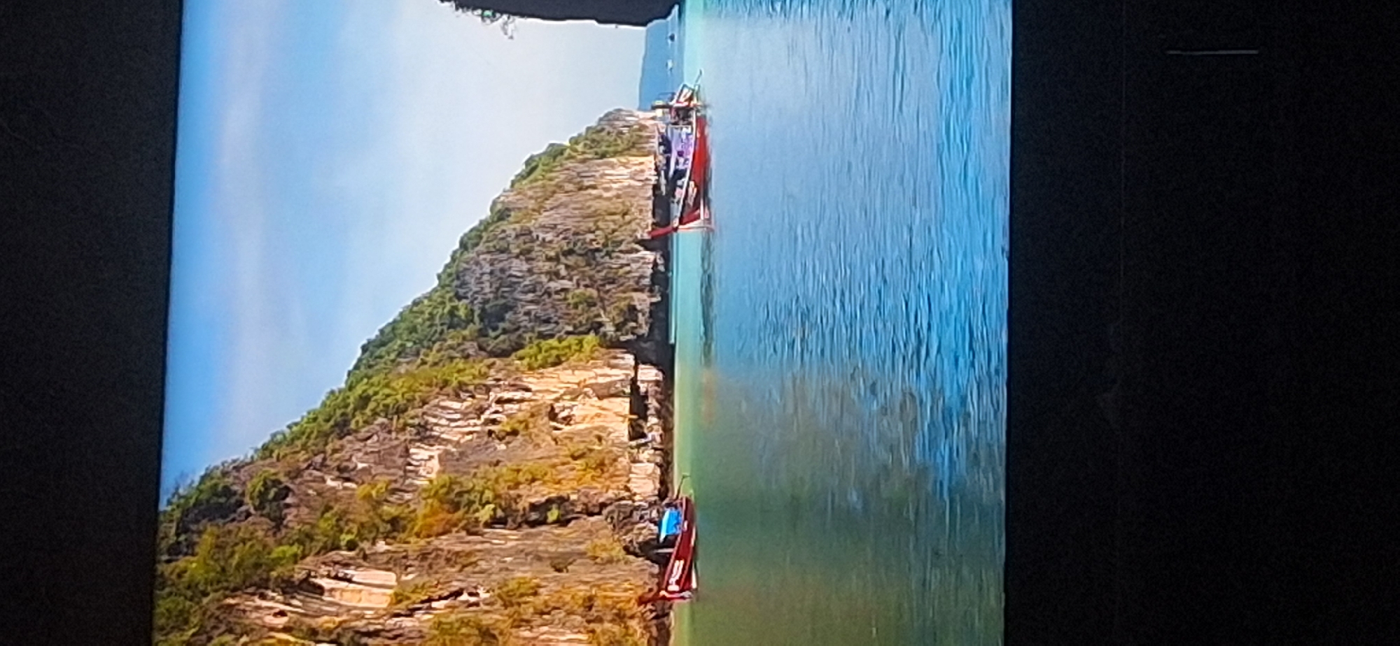The image depicts a scenic tropical coastline with steep, verdant cliffs rising from a turquoise sea. Two longtail boats, characteristic of Southeast Asia, are visible in the water. One boat, predominantly red, is closer to the shore, while another, also red with purple accents, is further out. The water is clear and calm, reflecting the bright blue sky. The cliffs are covered in lush green vegetation, with visible rock formations. The scene is peaceful and idyllic, suggesting a popular tourist destination. This image is not located in Bila Tserkva, Ukraine.