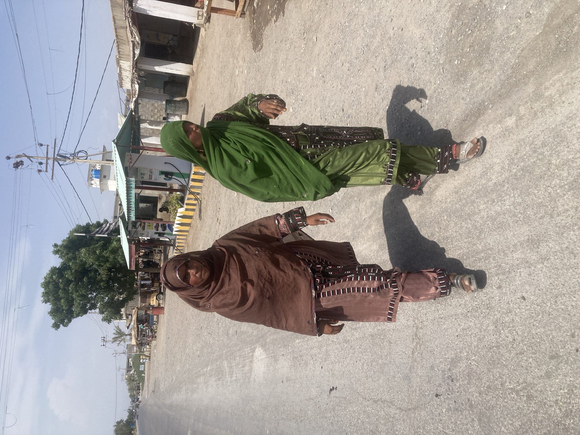 This media file captures an outdoor street scene in Uthal, Pakistan, depicting two women walking along a paved road under bright daylight.
**Subjects:**
The primary subjects are two women, positioned in the foreground and midground, walking towards the viewer's right. Both are dressed in traditional South Asian garments, specifically shalwar kameez, with their heads covered by dupattas. The woman closer to the foreground wears a brown outfit with a matching head covering. Her dress features intricate embroidery and mirror work, particularly visible on the sleeves, chest, and lower part of the kameez and shalwar. The second woman, slightly behind and to the left, is dressed in a vibrant green outfit with a matching green head covering, also adorned with elaborate embroidery and possibly mirror work on the fabric. Both women are wearing open-toed sandals or chappals. Their faces are partially visible.
**Scene and Setting:**
The setting is a street lined with low-rise buildings and simple structures, typical of a semi-urban or rural area. On the left side of the road, there are utility poles with a dense network of electrical or communication wires crisscrossing the sky. Further down, a large leafy tree stands out against the clear sky, and several smaller trees or bushes are also visible. There's a structure with a green corrugated roof on the left, possibly a small shop or commercial establishment, next to what appears to be a white machine or dispenser, possibly related to fuel or utilities, marked with yellow and black striped barriers. Buildings on both sides are simple, concrete or brick constructions. The road surface is a light grey, consistent with asphalt or concrete, showing some texture.
**Activity and Notable Details:**
The women appear to be walking casually along the road, going about their daily activities. The time of day is bright, indicating clear weather and sunshine, with strong shadows cast by the women and other objects, suggesting either mid-morning or mid-afternoon. The traditional attire with intricate embroidery is a significant cultural detail, likely indicative of Balochistani traditional dress. No discernible English text is visible in the image. The overall impression is that of a typical day in a local street in Uthal, Pakistan.
