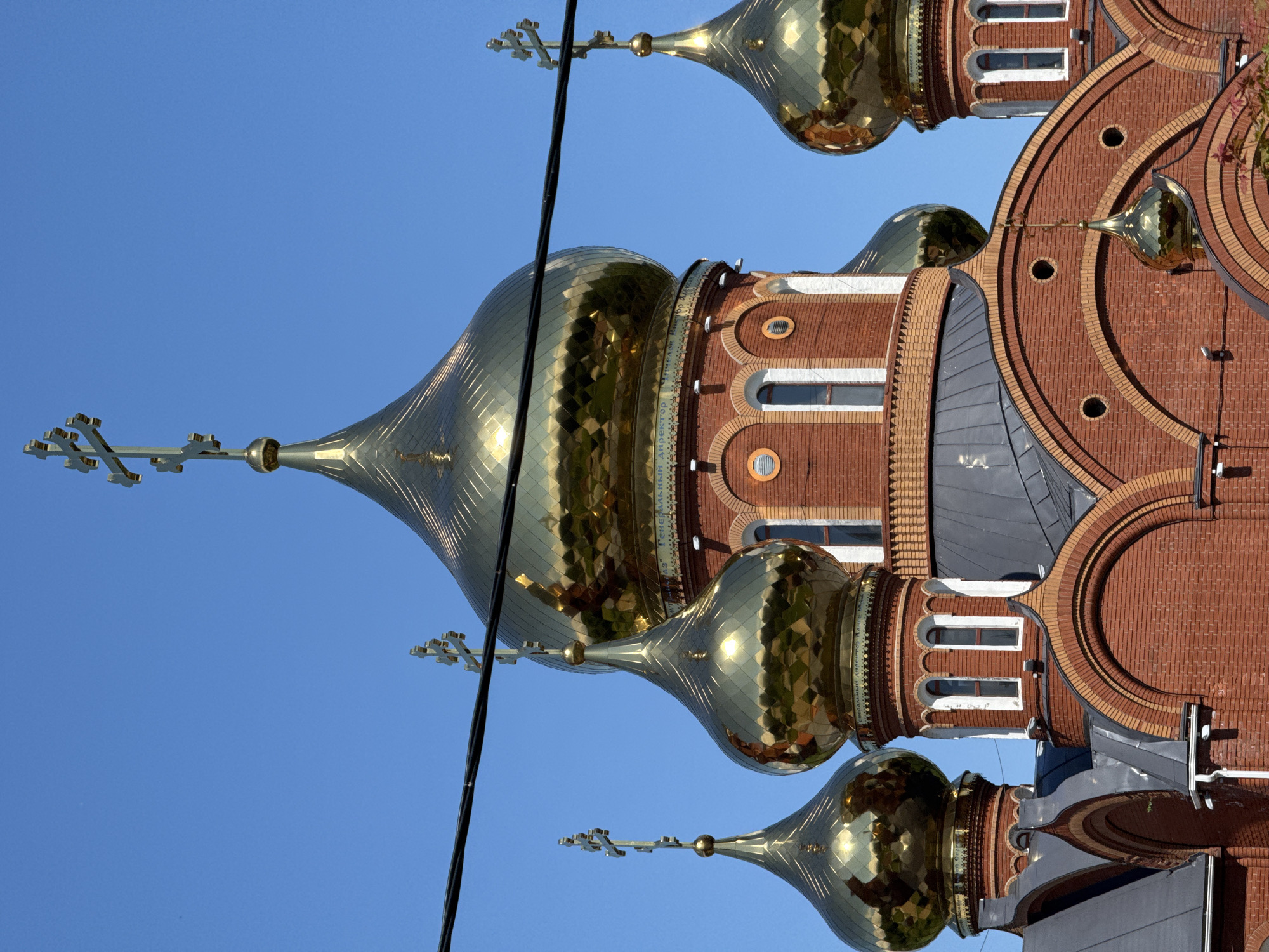A vibrant, sunlit photograph captures the upper sections of an Orthodox Christian church against a clear, bright blue sky. The image primarily features multiple golden, onion-shaped domes, each topped with an ornate Orthodox cross. These domes are highly reflective, showing numerous facets that glint brightly under the sun, suggesting a clear and sunny day.
The church building itself is constructed from reddish-brown brick, complemented by decorative lighter brickwork that forms intricate arches and patterns around windows and cornices. Several small, arched windows are visible on the brick structure. Dark grey or black roof sections are also discernible between the various brick elements and domes.
A thick, black cable runs diagonally across the upper-left portion of the frame, partially obscuring one of the larger golden domes and its cross. On a decorative band encircling the base of one of the larger domes, Cyrillic text is visible. The legible portion reads "ГЕНЕРАЛЬНЫЙ ДИРЕКТОР НИКОЛАЕВ Н. И." (General Director Nikolaev N. I.). The architecture is characteristic of Eastern Orthodox churches, situated in Chaikovsky, Russia. No people or specific activities are depicted; the image focuses solely on the architectural details of the building under bright daylight.