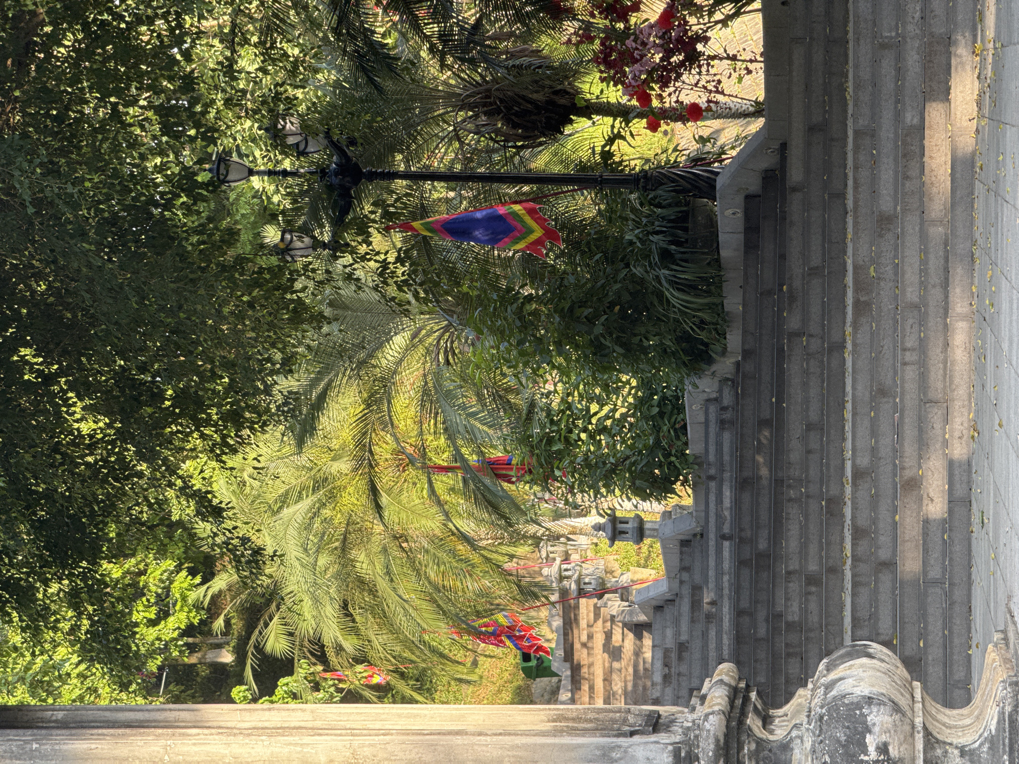 A sunlit outdoor scene, possibly in Lao Cai Ward, Vietnam, presents a downward view from an elevated vantage point, likely a stone staircase. The staircase descends alongside a stone wall. Lush greenery, including palm trees, fills the left side of the frame. A decorative street lamp stands next to the staircase, adorned with a colorful banner. Additional decorations, possibly flags or streamers, are strung between trees. The scene is bathed in natural light, creating a warm, vibrant atmosphere. No people are visible.