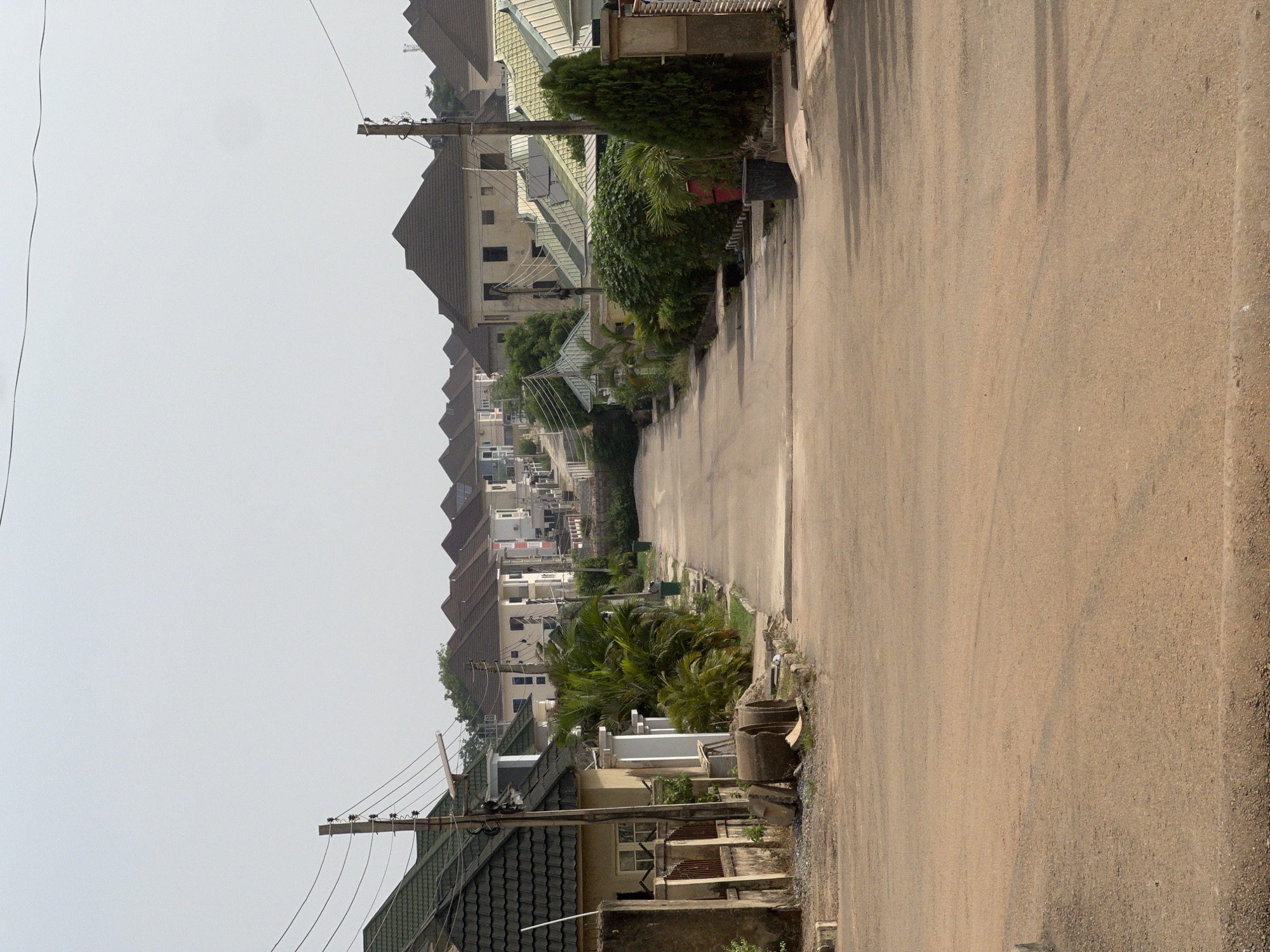 The image depicts a street in Abuja, Nigeria. A paved road stretches into the distance, flanked by buildings and vegetation. The buildings appear to be residential houses with dark-colored roofs. Electrical poles and wires are visible along the roadside. The sky is overcast, creating a diffused lighting condition. The road is empty of vehicles and people. The scene suggests a quiet, residential area.