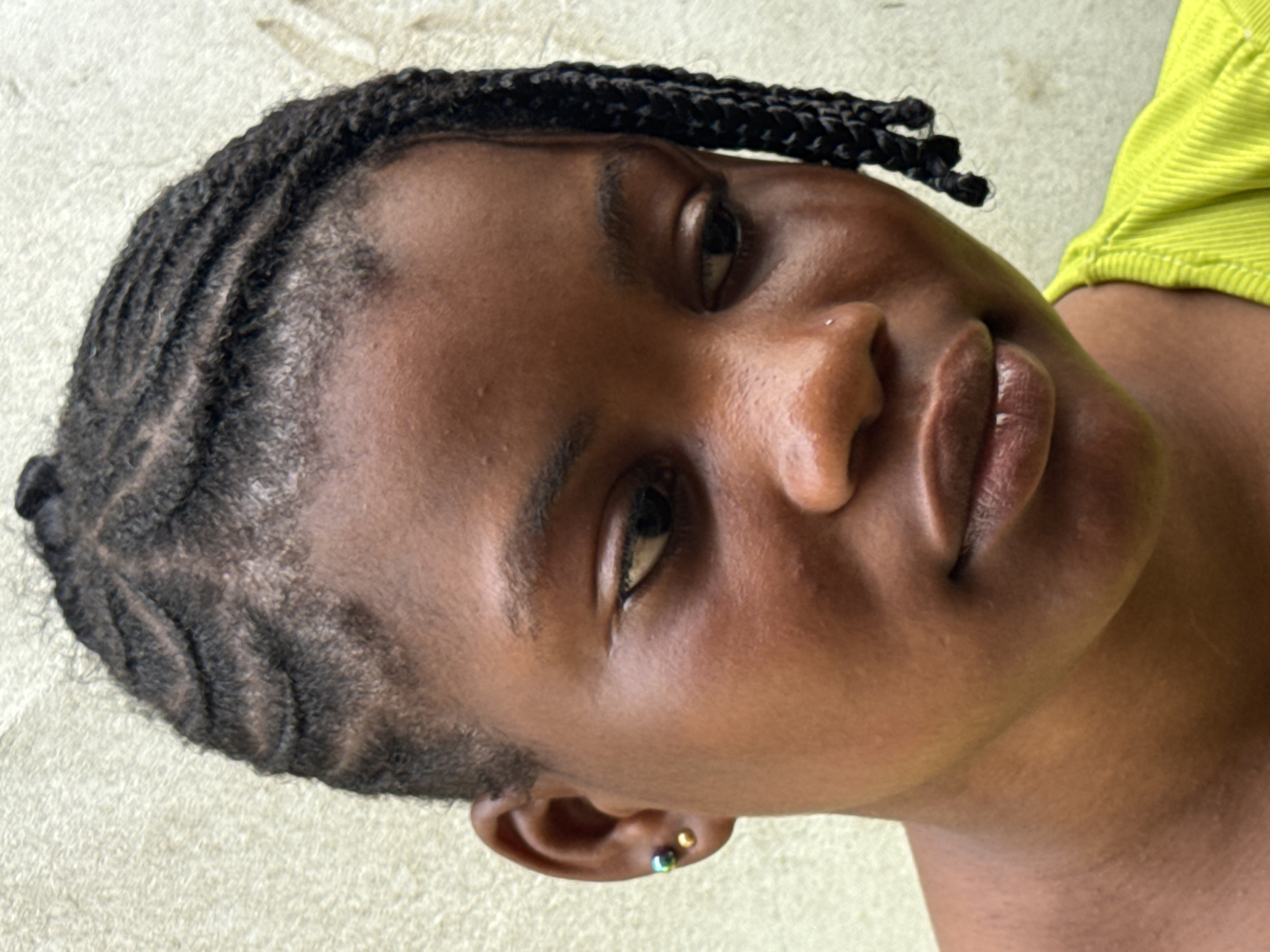 A close-up shot of a Black woman's face and upper body. Her hair is braided in cornrows that transition into small braids near her forehead. She is wearing earrings. A green shirt is visible on her right side. The background is nondescript, with no identifying features to indicate the location of Badagry, Nigeria.