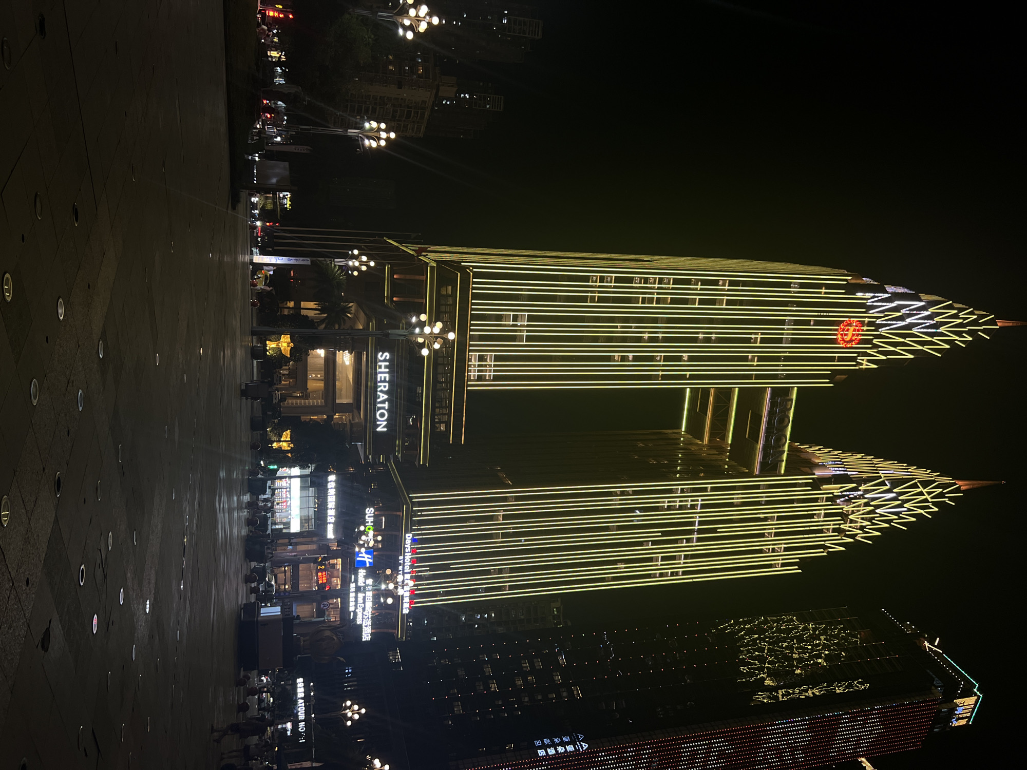 This is a nighttime, eye-level, outdoor photograph of the Sheraton hotel in Nan'an District, China. The main subject is the brightly lit Sheraton building, which is illuminated by numerous vertical yellow lights and features a prominent red Sheraton logo at its peak. The building has a unique architectural design with two towering structures connected by a bridge-like section. To the right of the Sheraton, another tall building is visible, also illuminated with various colored lights. In the foreground, a wide, dark, paved plaza is visible with scattered circular lights embedded in the ground. Several streetlights with multiple bulbs are positioned along the plaza. In the mid-ground, there are some trees and smaller buildings, also with lights on. A few individuals are present in the plaza, appearing as dark silhouettes against the illuminated background, with some walking and others standing. The sky is dark and clear, suggesting it is nighttime. The overall scene depicts an urban environment at night, with a focus on the illuminated architecture of the hotel and its surrounding plaza.