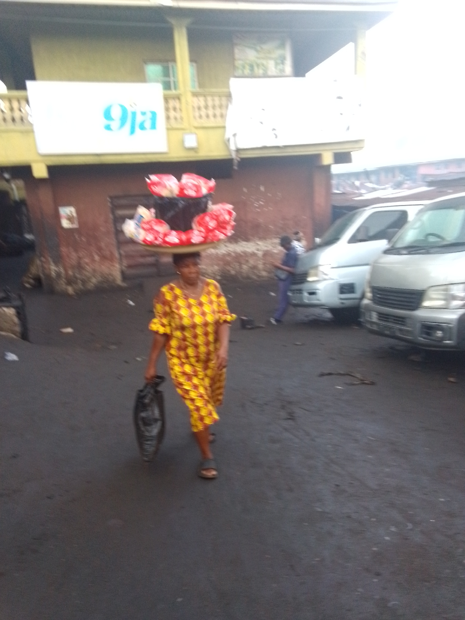 The image depicts a woman in Onitsha, Nigeria, carrying goods on her head and a black bag in her left hand. She is wearing a yellow and red patterned dress and sandals. She is walking on a dark, paved surface in what appears to be a market area. In the background, there are two light-colored vans parked along a building. A sign with "9ja" is visible on the building. Another person in purple pants and a white shirt is walking away from the scene, next to the vans. The overall setting suggests an urban street scene with commercial activity.
