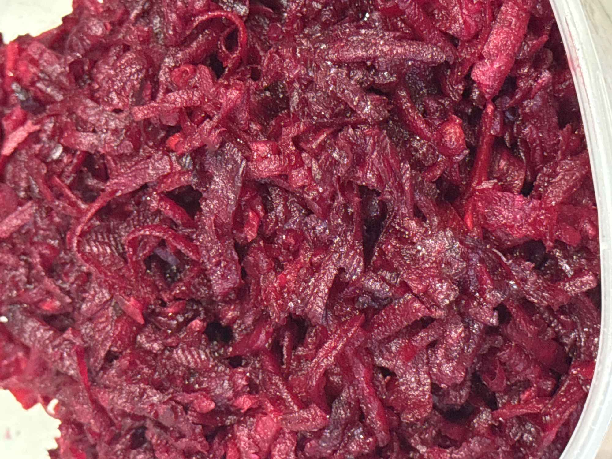 This is a close-up, overhead shot of shredded beets in a clear plastic container. The shredded beets are a deep red, almost purple, color and appear to be slightly glistening, possibly from a dressing or moisture. The texture is fine and stringy, with some pieces appearing thicker than others. The lighting suggests an indoor setting, with reflections visible on the shredded beets. There are no people or specific location cues in the image, other than the container itself, which could be found in a kitchen or grocery store. The focus is entirely on the shredded beets, suggesting they are the subject of interest, perhaps for a recipe or as a food item. There is no visible text in the image.