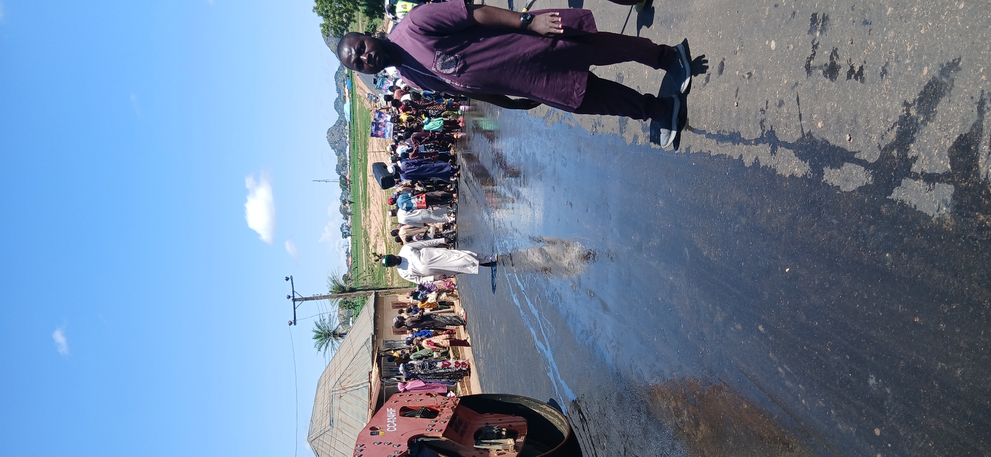 A group of people are gathered on a wet asphalt road, possibly in Kayarda, Nigeria, where roadwork is underway. A large steamroller is visible on the left side of the image, its red paint chipped in places and bearing the inscription "CC424HF". The wet road surface reflects the bright sky and the surrounding environment.
In the foreground on the right, a man in a deep purple traditional outfit with intricate embroidery on the chest is standing and looking directly at the camera. He is wearing grey athletic shoes. To his left, a man in a white flowing robe and a green cap is walking away from the camera, looking towards the crowd.
The crowd consists of individuals of various ages, many of them children, dressed in a mix of traditional and casual clothing. Some are observing the roadwork, while others appear to be interacting with each other. Several people are carrying items, including what appears to be a large black speaker. In the background, there are buildings, palm trees, and a utility pole. Further in the distance, rolling hills and a communication tower are visible under a clear blue sky with scattered clouds. The overall scene suggests a community event or a disruption caused by the road construction.