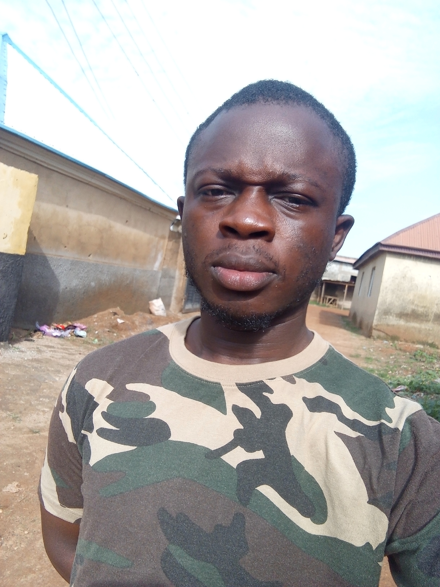 A medium shot, taken from a low angle, captures a young Black male looking directly at the camera. He has dark skin, short black hair, and a short beard. He is wearing a camouflage t-shirt. The background shows a dusty, dirt road with several buildings and a clear sky with power lines. The buildings appear to be simple, stucco structures, some with corrugated metal roofs, suggesting a rural or semi-urban environment. The overall scene has a candid, outdoor feel, possibly in a residential area of Abuja, Nigeria.