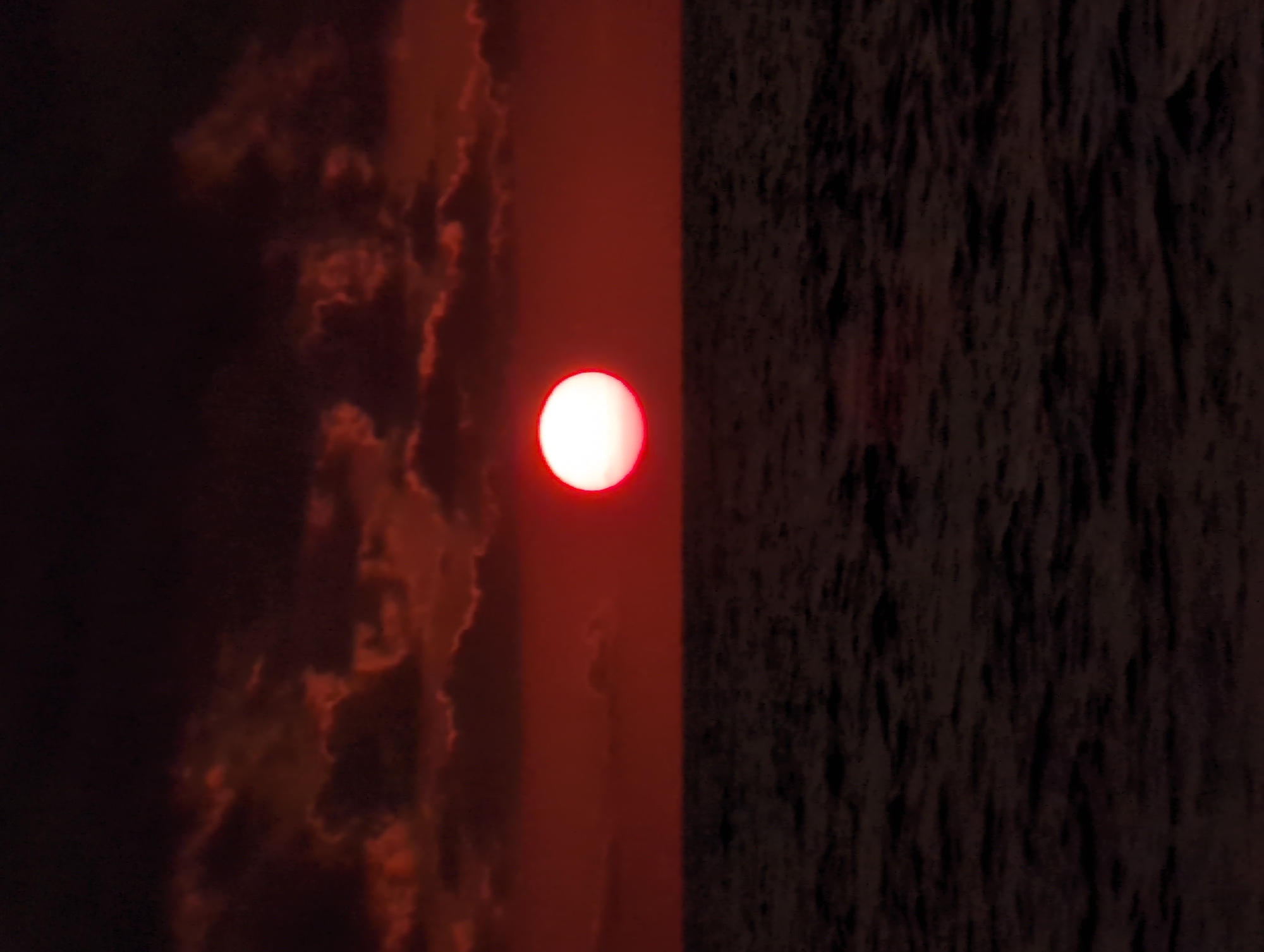 A dramatic sunset over the Black Sea casts a deep red hue across the sky and ocean. The sun, a bright white orb with a red halo, is positioned just above the horizon line. Dark, ominous clouds, also tinged with red, fill the upper portion of the frame. The sea is dark and choppy, with subtle ripples reflecting the dim light. There are no people or man-made objects visible in the image, creating a natural and serene, yet intense, atmosphere. This scene is likely to be observed from the coast of Shekvetili, Georgia, a region known for its natural beauty and coastal views.