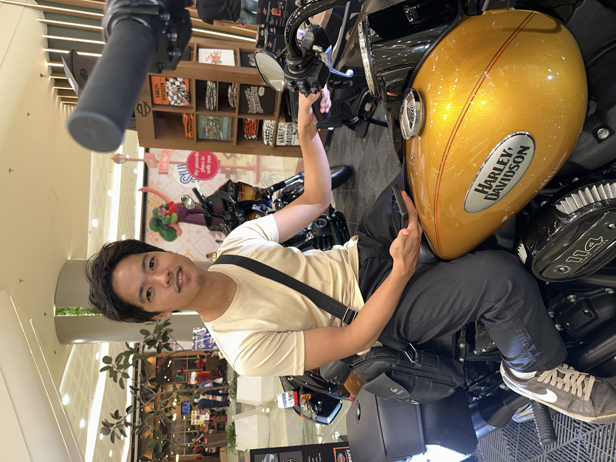 A young Filipino male is seated on a Harley-Davidson motorcycle inside a shopping mall in Davao City, Philippines. He is wearing a beige t-shirt, black pants, and athletic shoes. A black crossbody bag is slung over his shoulder. He holds a smartphone in his left hand and is reaching out with his right hand towards the motorcycle's handlebars. The motorcycle is a prominent golden color with "Harley-Davidson" branding on the fuel tank. Behind him, shelves display Harley-Davidson merchandise, including t-shirts and accessories. In the background, other people are visible, along with mall decorations and structural elements like pillars and ceiling lights. The environment appears to be a retail space within a modern shopping complex.