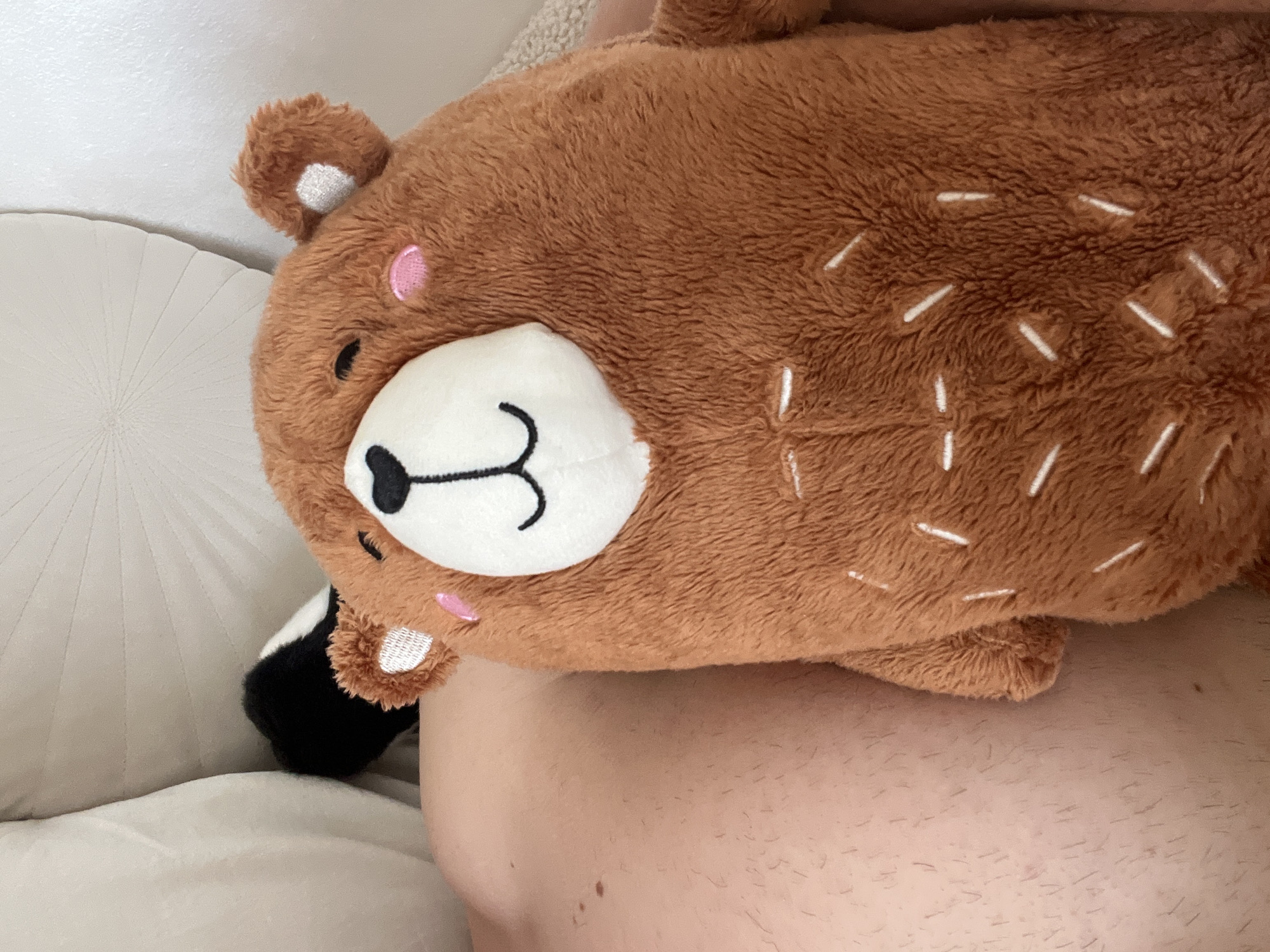 A brown stuffed bear toy is resting on a person's bare stomach. The bear has white spots on its brown fur, a white face with a black nose and mouth, and pink circles on its cheeks. It is lying on what appears to be a white pillow or bed. The person's bare stomach is visible beneath the bear. The photo was not necessarily taken in Blagoevgrad, Bulgaria.