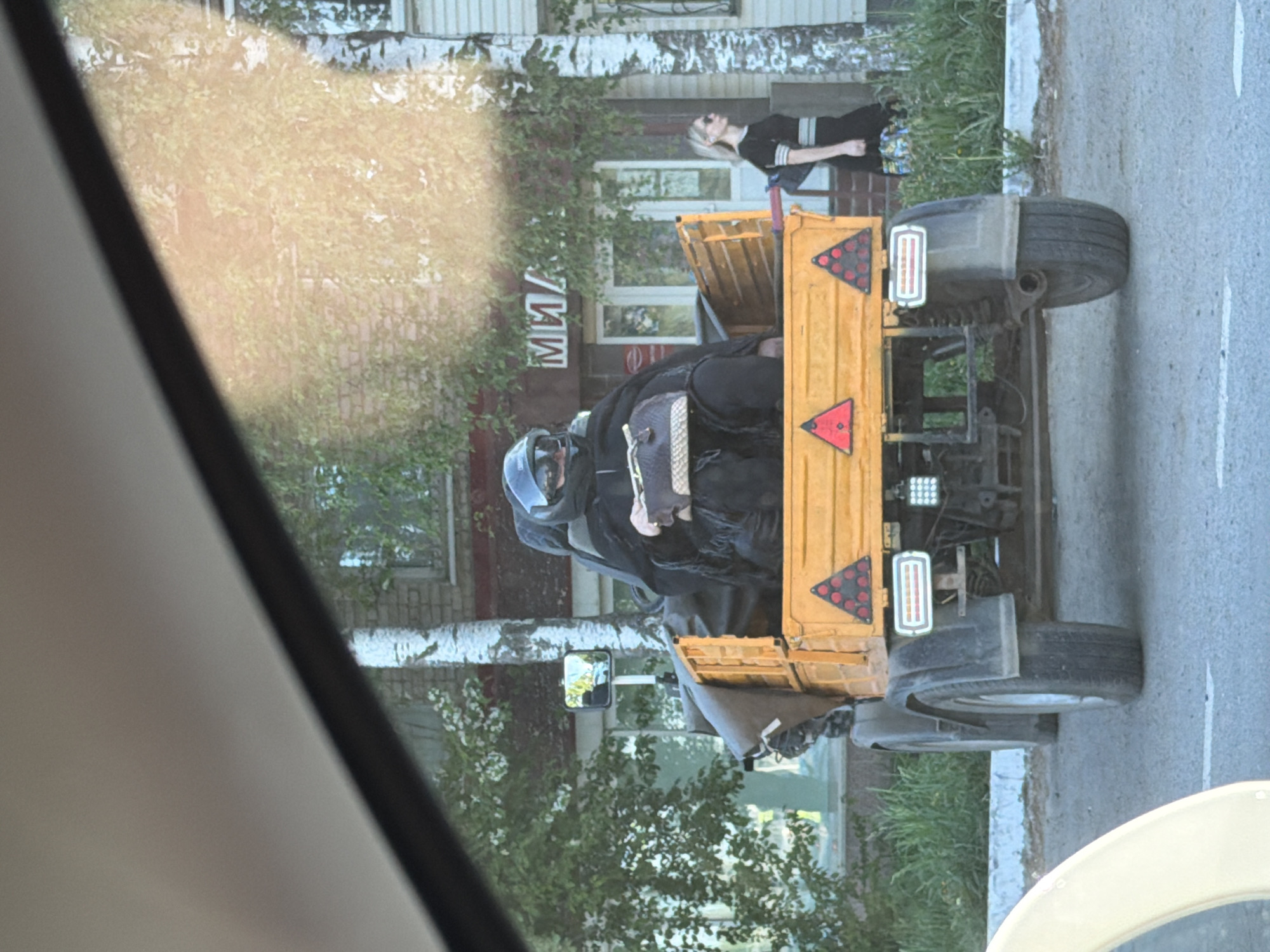 A photograph, taken through a vehicle window, shows a yellow small utility vehicle with a driver wearing a helmet and protective clothing. The vehicle appears to be a modified agricultural vehicle, with rear-mounted signal triangles and LED lights. A woman is leaning against a building in the background. The location is Chaikovsky, Russia, indicated by the Cyrillic text “МИЛ” on the building. The image is cropped on the left by the vehicle window frame.