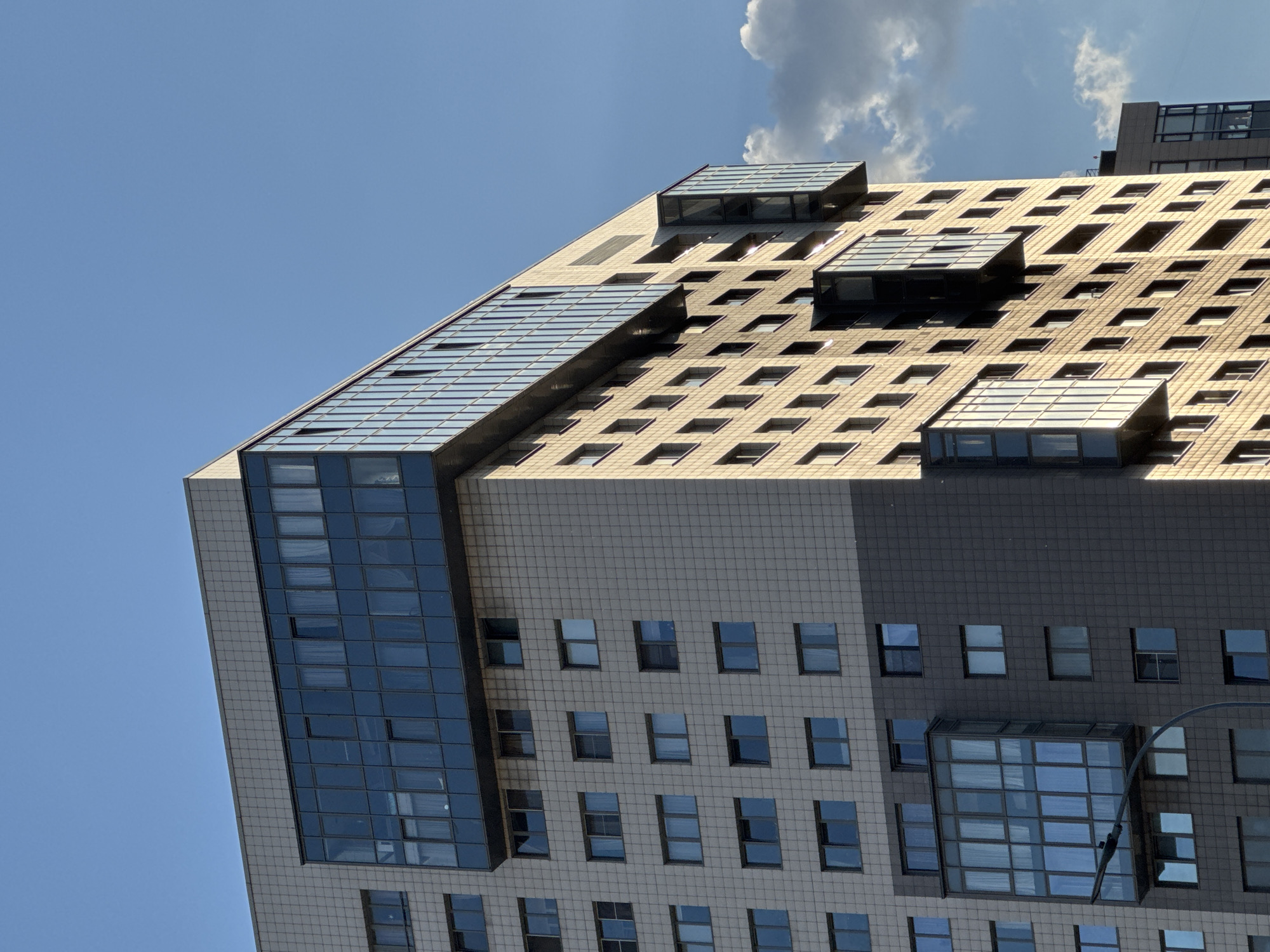 An upward-angled photograph captures a modern, multi-story building against a clear blue sky with one prominent white cloud in the upper right. The building features a complex geometric design, characterized by contrasting sections of light grey and dark grey paneled facades, interspersed with numerous square and rectangular windows.
The upper and right sides of the building are dominated by large, angled glass structures that appear to be skylights or sloped curtain walls. These glass elements, reflecting the bright blue sky, have a distinct grid pattern and some protrude from the main facade, adding to the building's dynamic shape. The bright sun, likely late morning or early afternoon, illuminates parts of the building, casting sharp shadows that emphasize the architectural details and textures. The light grey sections of the facade appear warm and yellowish where the sun hits directly, while the dark grey sections are partially in shadow. The lower part of the building shows more conventional window arrangements within the grey paneled walls, with some recessed sections incorporating additional glass elements. There are no people, visible activities, or discernible text in the image, focusing solely on the architectural structure under clear, sunny weather.