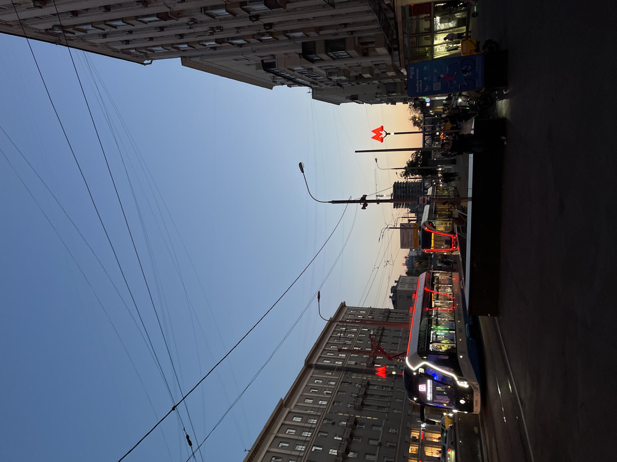 The image depicts a street scene in Moscow, Russia, at dusk. Two trams are visible on tram tracks. The tram in the foreground is modern, with illuminated windows and red and white exterior lights, displaying "T2" on its front. The second tram, further down the track, also shows interior lights. Several buildings line the street, with windows reflecting the evening sky. Overhead power lines are prominent. Two red metro signs, a distinctive "M" symbol, are visible. People are present on the sidewalks, some standing, and a few are near a sign that appears to be advertising a financial service. Electric scooters are parked along the sidewalk. The sky is a gradient of blue, transitioning to a soft orange and pink at the horizon, indicating sunset. The lighting suggests the time is shortly after sunset.