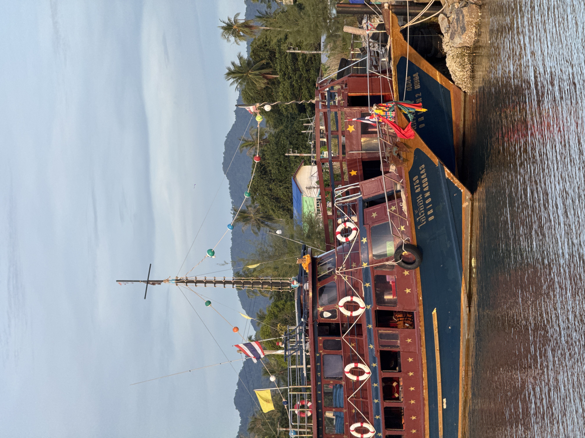 Two maroon and blue tour boats are moored side-by-side in a harbor in Ko Pha Ngan, Thailand. The boats are decorated with yellow stars and life preservers. The boat on the left has Thai writing and numbers "4750" on its hull, while the boat on the right has Thai writing and numbers "01504" on its hull. Mountains and palm trees are visible in the background. The sky is cloudy and the water is calm.