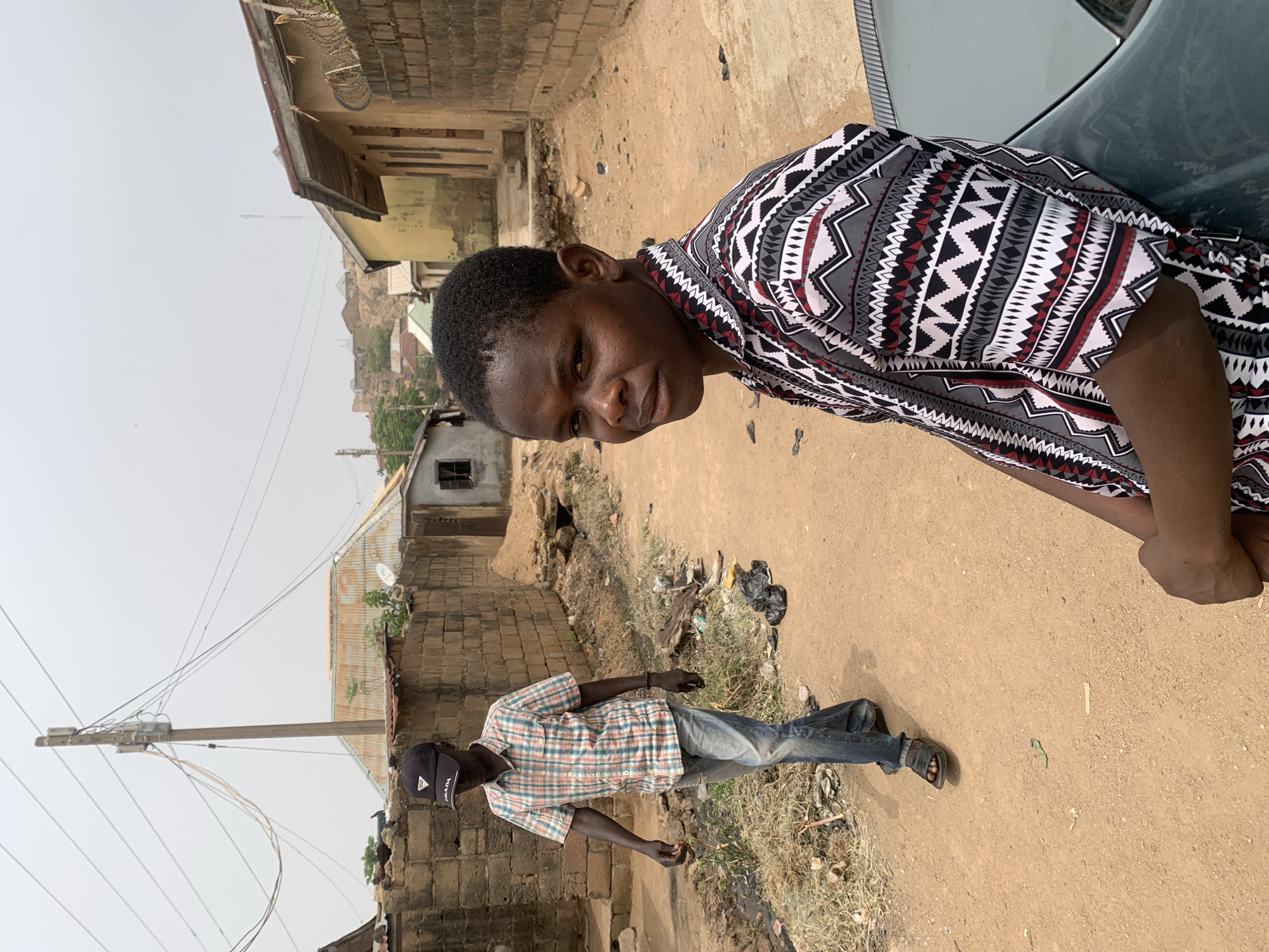 The image depicts an outdoor scene in what appears to be a residential area. In the foreground, a person with dark hair and skin is leaning towards the camera, wearing a patterned shirt with geometric shapes in black, white, and red. The person is positioned next to a car with a dark green or grey surface visible.
In the background, another individual is walking away from the camera down a dirt road. This person is wearing a plaid shirt, blue jeans, a black baseball cap and sandals. The road is flanked by brick and corrugated metal buildings, with visible signs of wear and tear.
Beyond the buildings, there are hints of more structures and terrain, indicating the presence of a town or city. The overall impression is of a modest, possibly impoverished neighborhood. The scene is lit by natural daylight, suggesting an overcast or hazy day. The location is Jos, Nigeria, as specified.