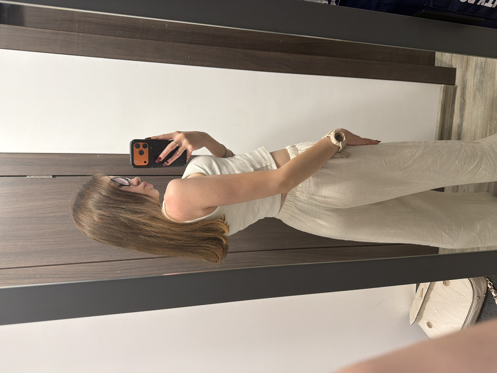 A young woman with long brown hair and glasses stands in front of a full-length mirror, taking a selfie with her phone. She is wearing a white tank top and beige linen pants. A white quilted handbag with gold accents is visible on a surface to her left. The scene appears to be an indoor changing room or dressing area, with wooden paneling and a white wall. The background suggests a residential or commercial setting, potentially a boutique or apartment. The lighting is even and natural. The woman is angled away from the camera, showing her profile and back. Her fingernails are painted a dark red. The overall impression is a candid moment of self-documentation.