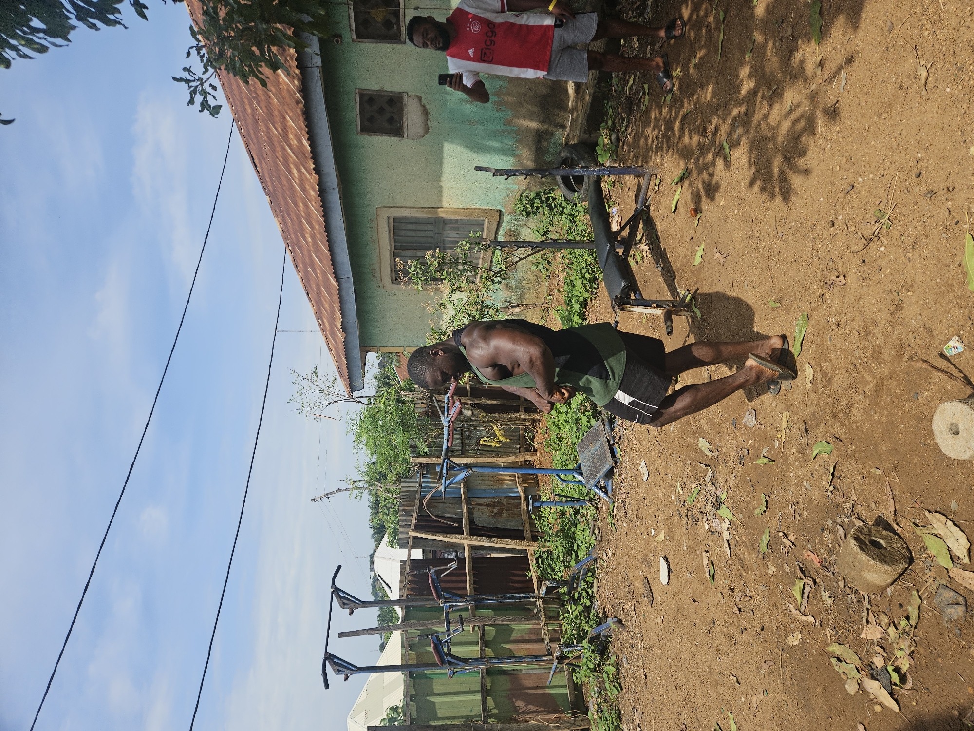 The image depicts a man in Akwanga, Nigeria, engaged in physical exercise outdoors. He is wearing a green tank top and black shorts with white stripes, and flip-flops. He's in the middle of a workout station. To his left, there are several other fitness apparatuses, appearing to be homemade from metal pipes, arranged near a corrugated metal fence. Another man, wearing a red and white Ajax jersey and grey shorts, stands by a building with a rusty metal roof, holding a black smartphone, possibly recording the first man. The ground is dirt, scattered with leaves and small objects, and a concrete weight rests on the right side of the frame. The area is partially shaded by trees.