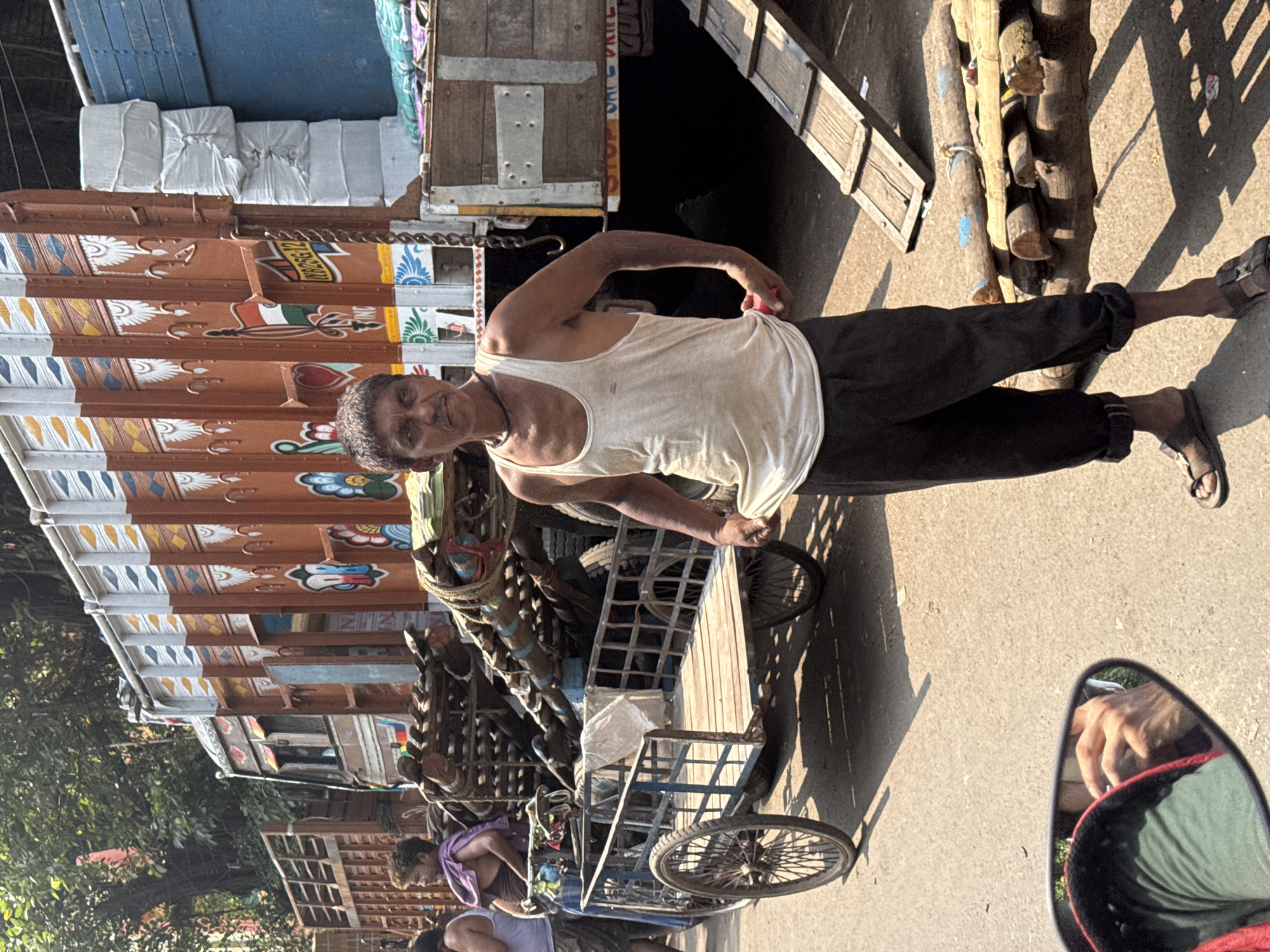 The image depicts a middle-aged man in Kolkata, India, wearing a white tank top and dark pants, standing next to a hand-pulled cart. The cart, made of metal and wood, is loaded with wooden logs. The man is partially obscured by the cart and a large, decorated truck parked on the street. The environment is a sunny, urban street. A side mirror reflects a hand and vehicle detail.