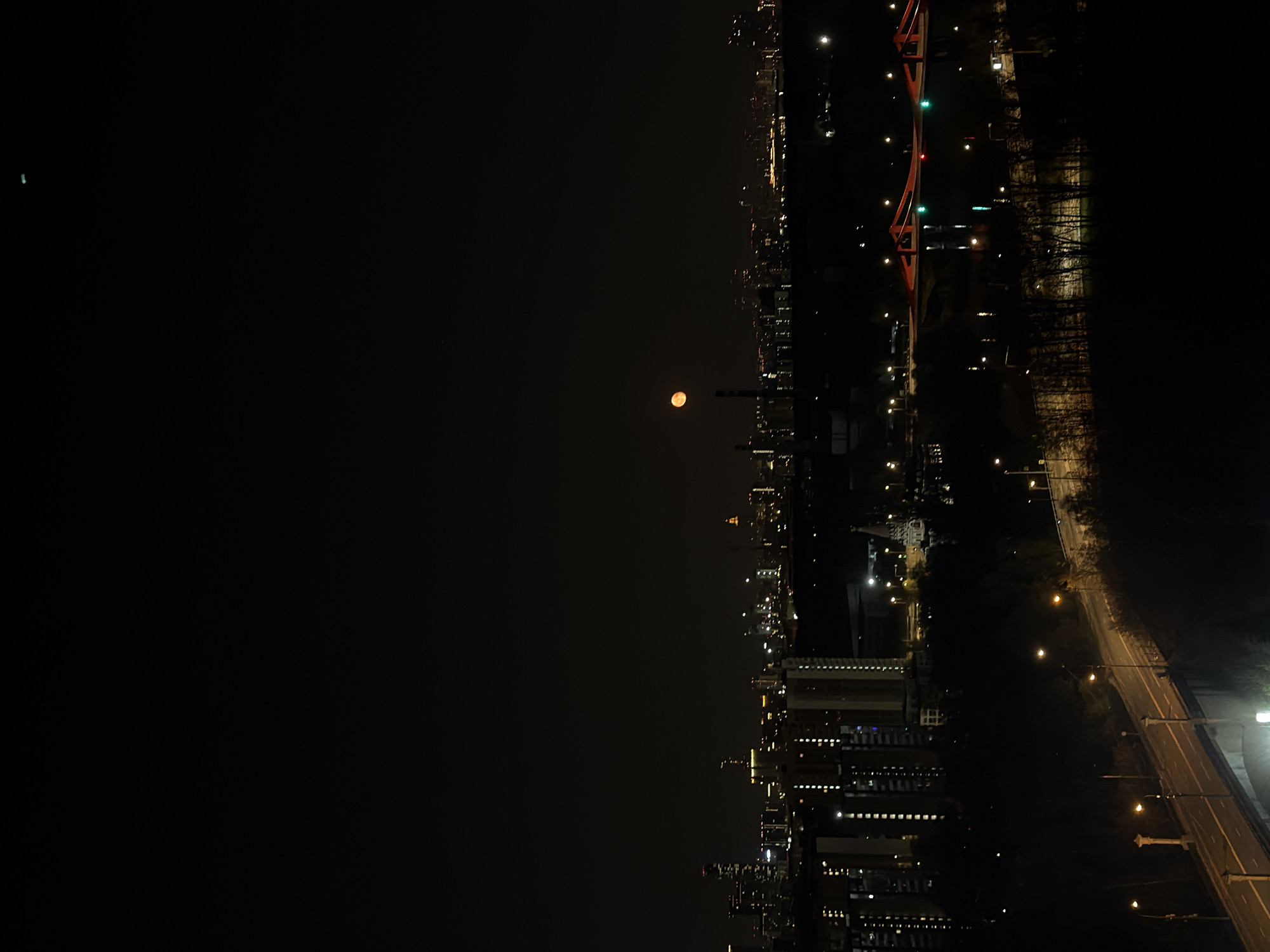 The image depicts a nighttime scene in Khimki, Russia, with a full moon visible in the dark sky. The city skyline is illuminated with lights from buildings and street lamps. A river runs through the city, with a bridge visible, marked by red and green lights. The road is lined with street lights. The environment is dark with city lights providing the only illumination. There are no people visible.