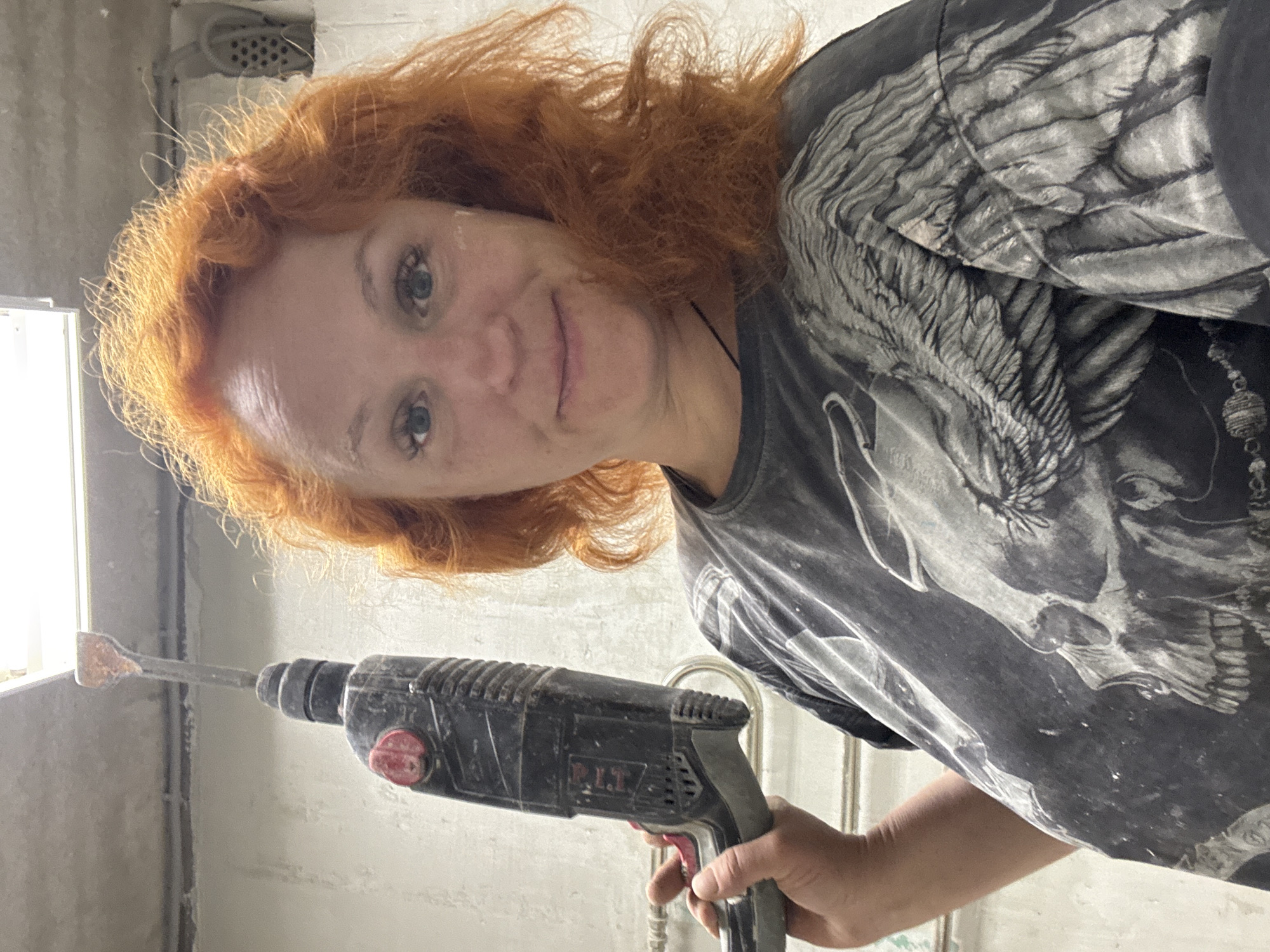 A red-haired woman is holding a black jackhammer in front of a light fixture. The jackhammer has a flat chisel bit. The woman is wearing a gray t-shirt with a skull and wing design. The background appears to be a concrete wall in a room under construction. The image was taken in Chaikovsky, Russia.