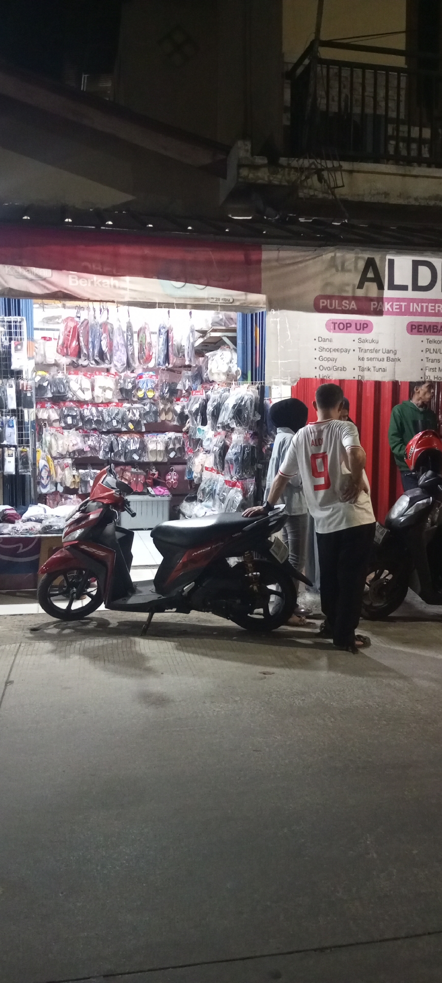 This is a medium-shot, eye-level, outdoor photograph taken at night in front of a storefront on a street in Setu, Indonesia. The storefront appears to be selling various accessories and goods. A red and black scooter is parked on the sidewalk in the foreground, angled to the left. The scooter's kickstand is down, and its headlights are off. The storefront is illuminated by artificial lighting, revealing shelves stocked with numerous items, many of which are packaged in plastic. Some items are hung from the ceiling. A banner with Indonesian text and logos is visible above the merchandise. To the right of the storefront, there's a larger black scooter. Three individuals are present: one person wearing a white t-shirt with the number "9" on the back and black pants, another person partially obscured by the first, wearing a light-colored outfit, and a third person in the background wearing a green jacket. The ground in front of the storefront is paved with concrete. The overall atmosphere is that of a small retail business operating at night.