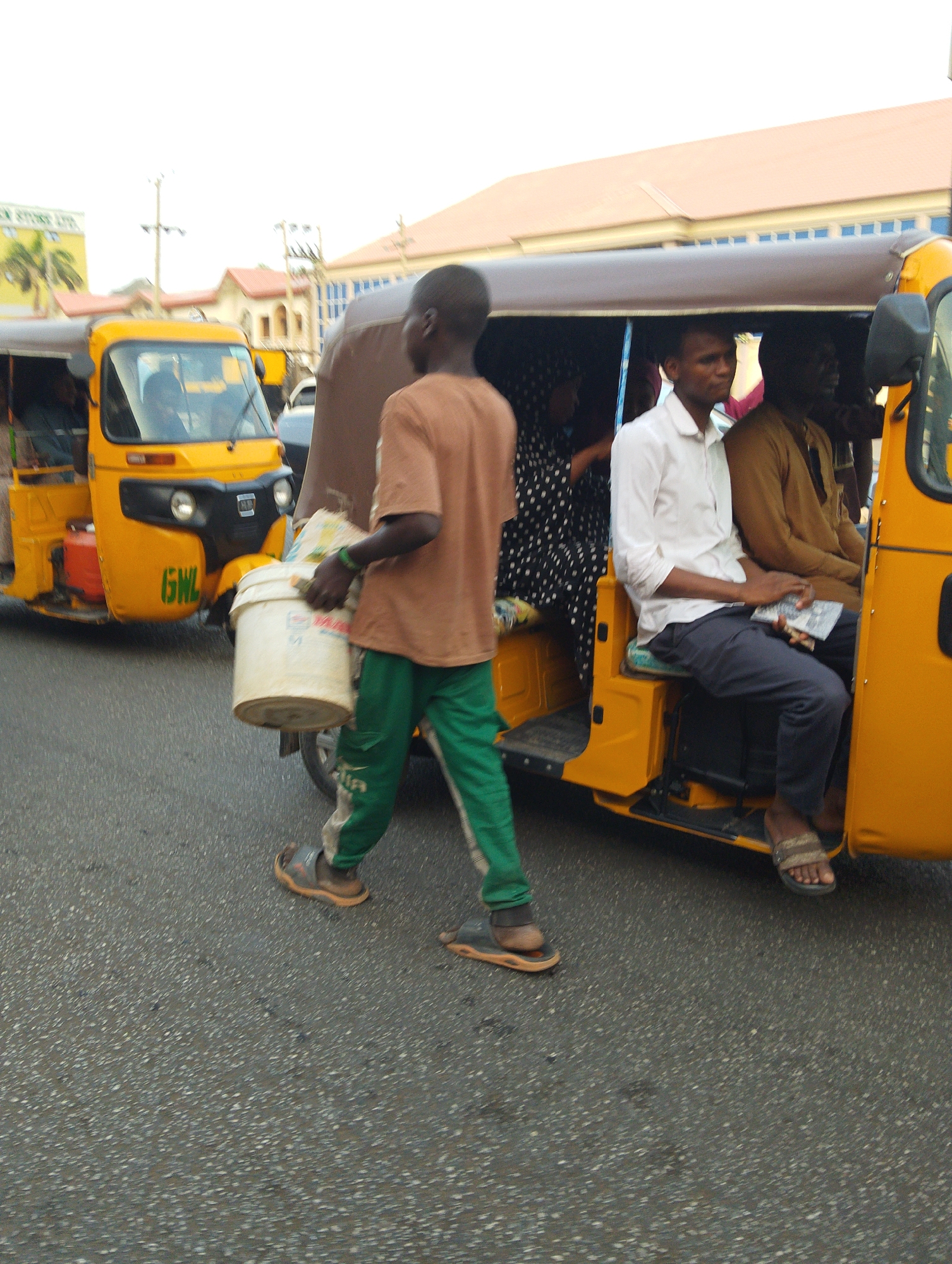 This image captures a bustling street scene in Babban Giji, Nigeria, featuring people and vehicles engaged in daily activities.
In the foreground, a young man walks from left to right, carrying a large white bucket in his left hand and a white plastic bag in his right. He wears a short-sleeved brown t-shirt, green sweatpants with white stripes, and dark sandals. A green wristband is visible on his left wrist. He appears to be a vendor or someone transporting goods.
To the right, a bright yellow motorized tricycle, commonly known as a Keke Napep, is either stationary or moving slowly. Several passengers are seated inside. A man in a white collared shirt and dark pants is prominent, holding what appears to be money in his lap. Next to him, another man in a brown garment is visible. Further inside, a person wearing a black and white polka dot outfit can be seen. The tricycle has a dark brown fabric canopy.
Another yellow Keke Napep is partially visible further down the street to the left, with the letters "GNL" painted in black on its front panel. The road surface is asphalt, speckled with light-colored debris.
In the background, under a bright, overcast sky, several multi-story buildings with reddish-brown roofs line the street. A distant sign, partially visible on the left, reads "STORE LTD" or similar. The scene depicts a typical urban environment during daytime, with people going about their routines, and the tricycles serving as common modes of transport. No strong emotions are discernible on the faces of the individuals; they appear focused on their immediate activities.
