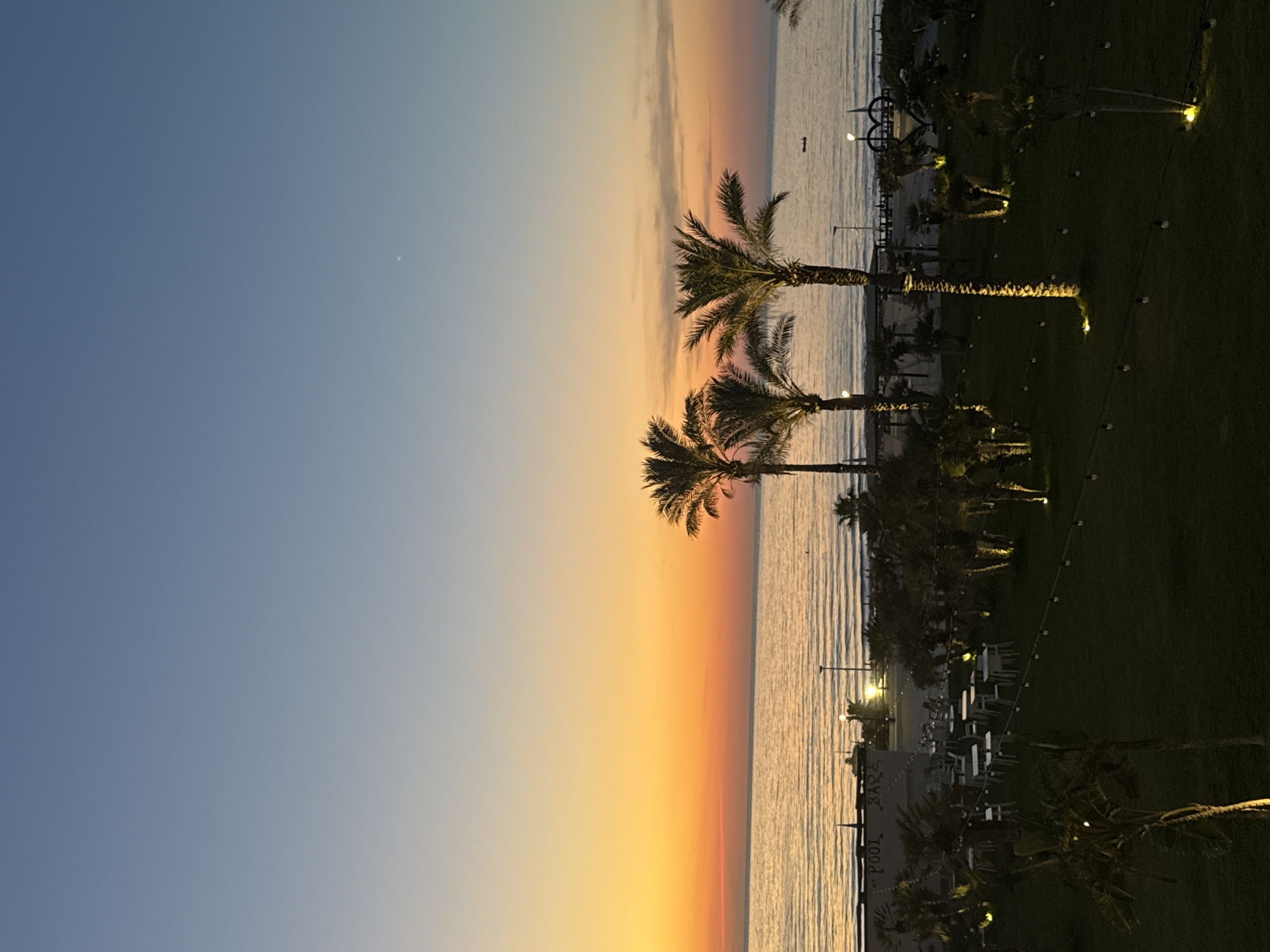 A vertical, eye-level, medium shot captures a tranquil sunset scene at a beach resort in Isola delle Femmine, Italy. The sky transitions from a pale blue at the top to a vibrant gradient of orange and yellow near the horizon, with the sun having just set below the sea. The calm, reflective ocean stretches across the middle of the frame, meeting the dusky sky. In the foreground, a lush green lawn is dotted with scattered white lights and several palm trees, their trunks illuminated by uplighting.
To the left, in the mid-ground, a building with "POOL BAR" signage is visible, with rows of empty white tables and chairs set up on a paved area, suggesting a dining or lounge space. The beach itself is visible beyond the lawn, lined with lounge chairs and umbrellas. Several more palm trees are scattered throughout the scene, some more illuminated than others. A small, dark boat is a distant speck on the water. The overall atmosphere is peaceful and serene, typical of a seaside resort at dusk.
