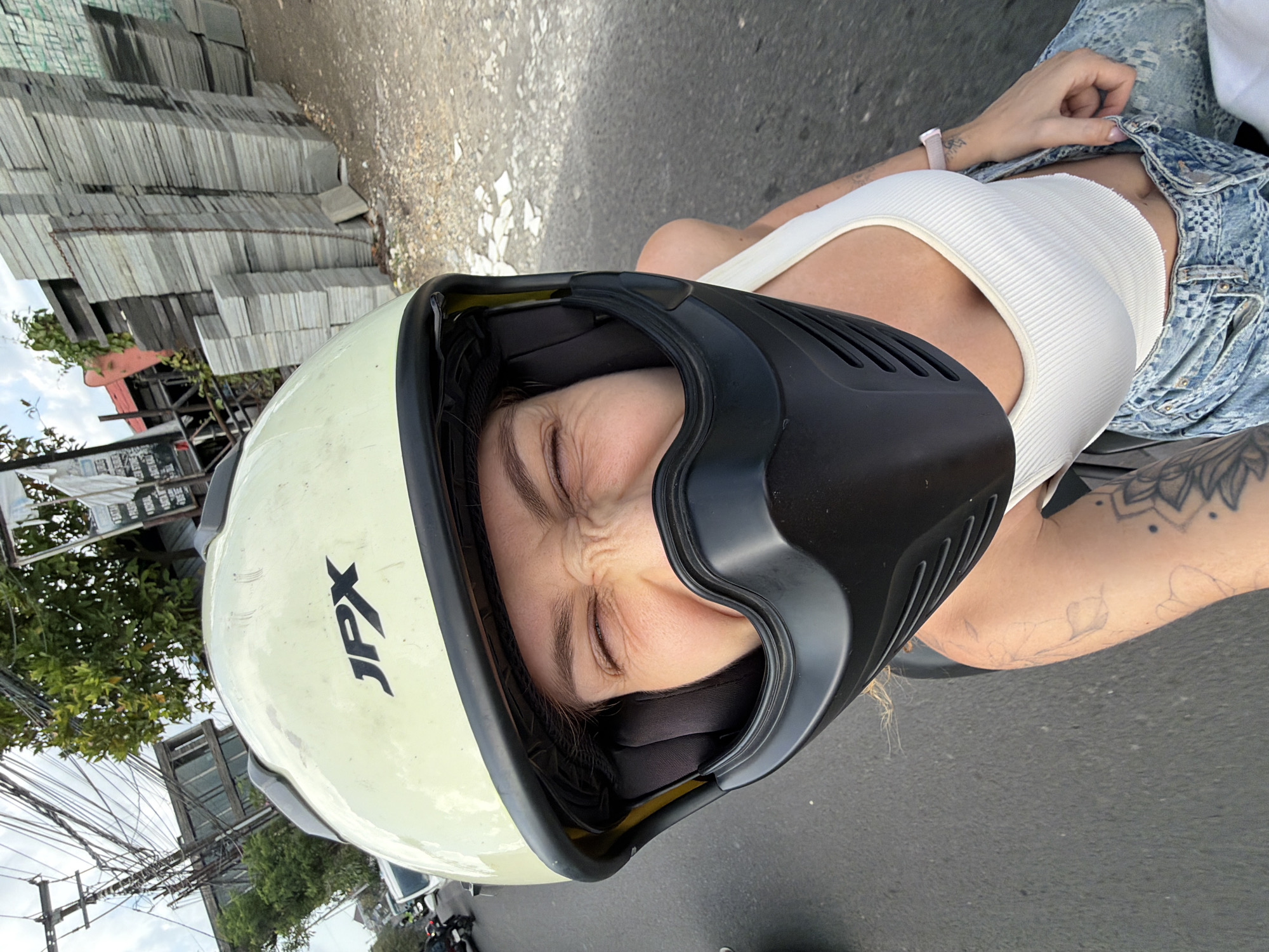 A person wearing a pale yellow full-face motorcycle helmet with a black visor and a white tank top is riding a motorcycle on a paved road in Mengwi, Indonesia. The person's eyes are squinted, and their face is partially obscured by the helmet. They are wearing blue jeans and have tattoos on their left arm and leg. The background shows a street scene with trees, power lines, buildings, and stacks of gray paving stones. The sky is cloudy.