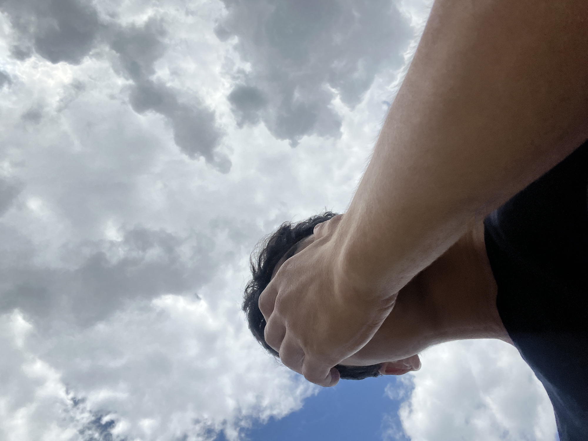 A low-angle, medium shot captures a person's head and arm from behind, looking up towards a cloudy sky. The person is wearing a black t-shirt and has dark hair. Their right arm is raised, with their hand covering their face and forehead. The background consists of a mix of blue sky and large, gray, cumulus clouds. The lighting suggests an overcast day. The image is taken outdoors, with no identifiable urban or natural landmarks, making it impossible to ascertain the location.