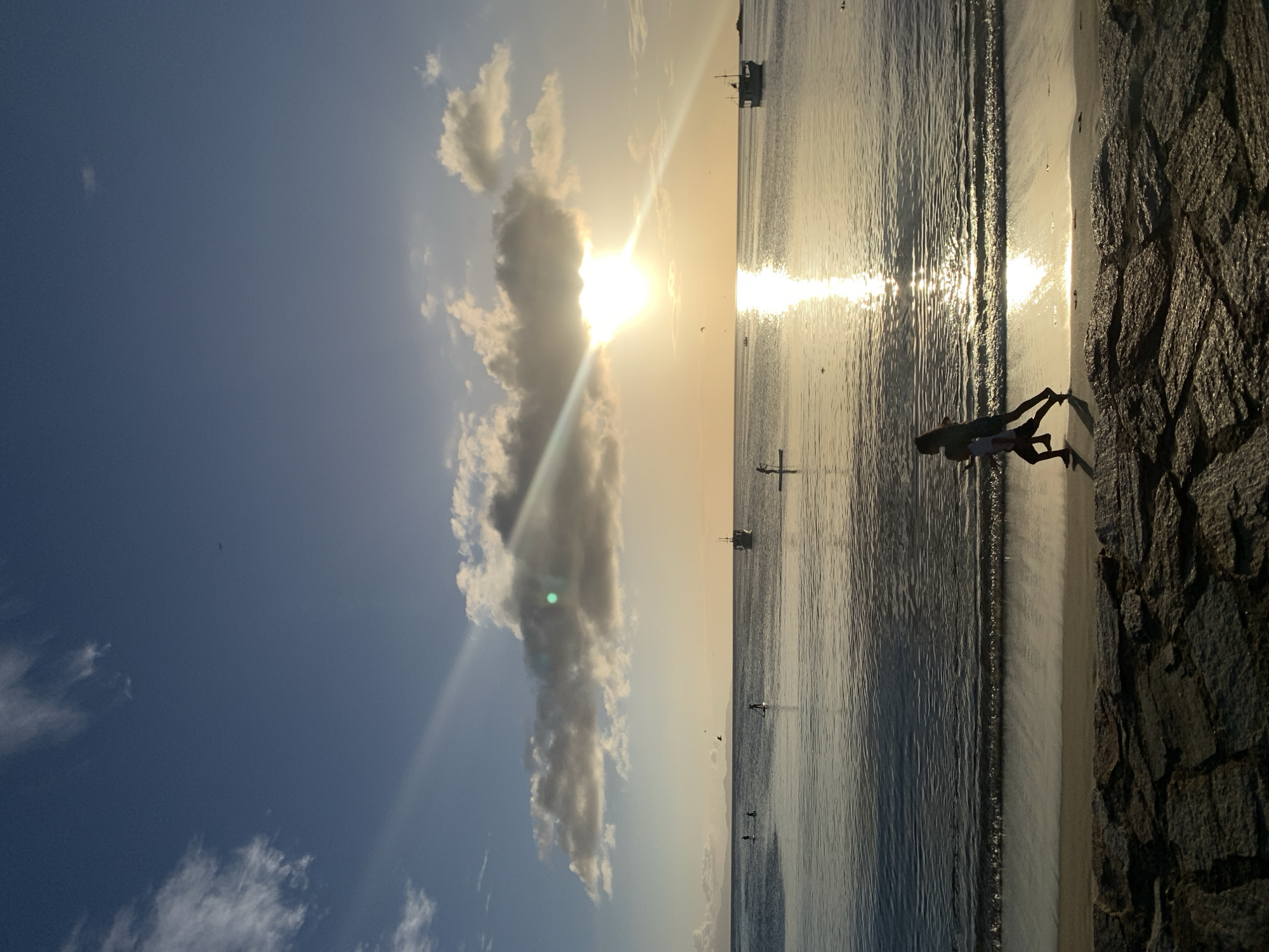 The image depicts a serene beach scene at sunset in Juan Griego, Venezuela. The sun, partially obscured by clouds, casts a warm glow across the calm ocean waters, creating a shimmering reflection. The sky is a blend of blue and orange hues, with scattered white clouds. In the foreground, a rocky embankment lines the sandy beach. Two figures, silhouetted against the setting sun, are seen running along the shoreline. Further out in the water, several individuals are paddleboarding, and a small boat is visible in the distance. The overall atmosphere is peaceful and tranquil, capturing a moment of natural beauty.