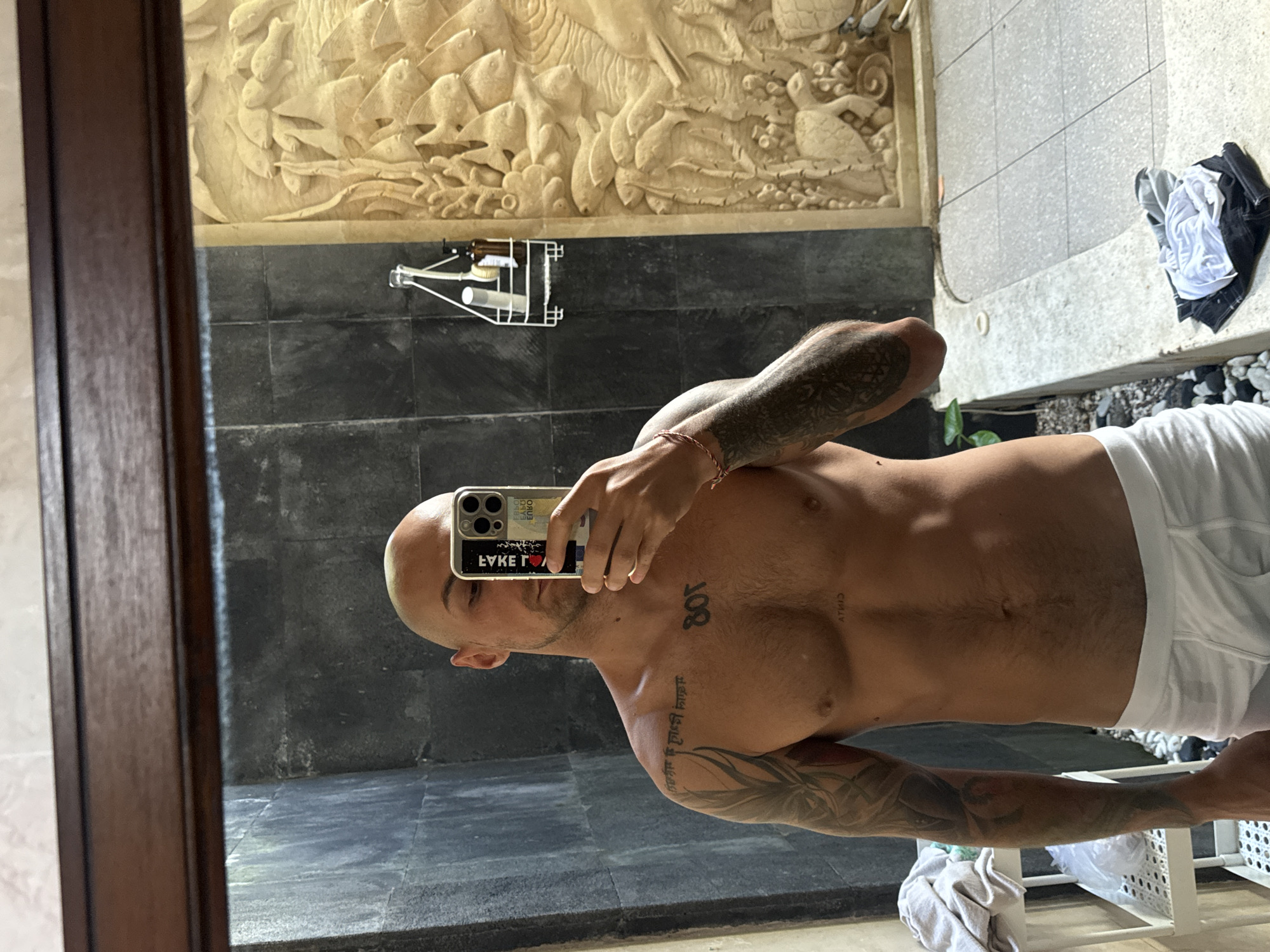 A bald man, wearing white boxer briefs, stands in front of a mirror in what appears to be an outdoor shower area. He holds a smartphone in his right hand, taking a mirror selfie. The man has tattoos on his left arm and chest, with the number "801" tattooed on his chest. Behind him, the wall is covered in dark, textured tiles, and to the right, a carved stone relief depicts marine life, including fish and turtles. A shelf with toiletries is visible on the wall. The floor is tiled, and there are some rocks and plants in the background. The environment suggests a tropical setting, likely a bathroom or outdoor living space in Kuta Selatan, Indonesia, known for its resorts and villas.