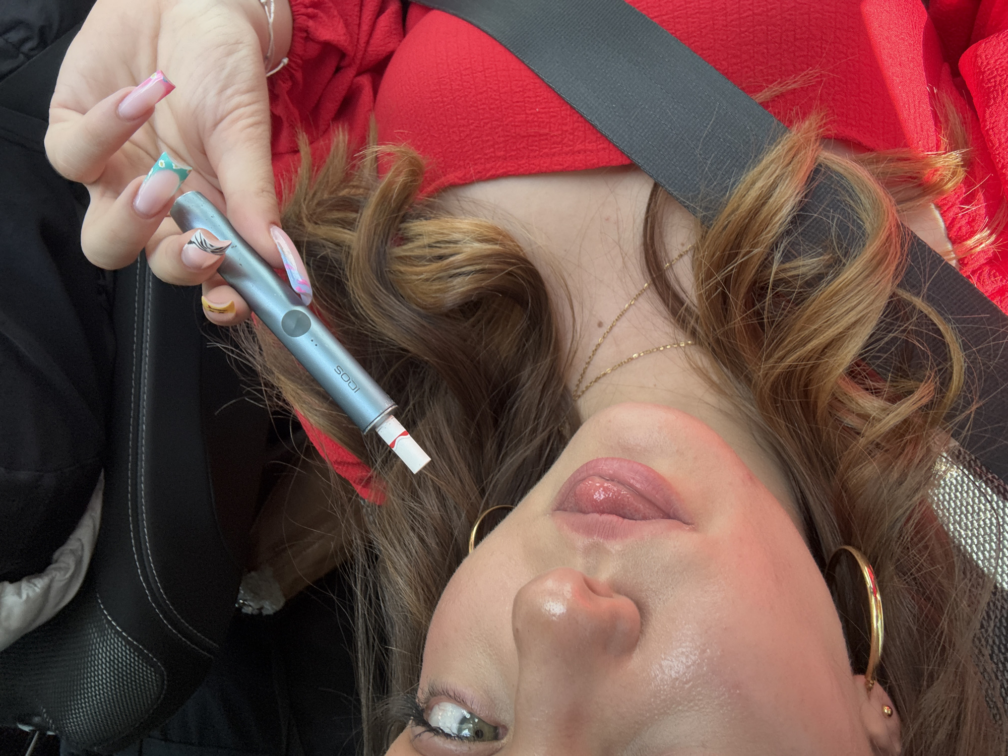 A selfie image of a woman in a car, wearing a red top and a seatbelt. She is holding a blue smoking device in her left hand, which has long, decorated nails. She is wearing gold hoop earrings and a gold necklace. The image is taken at an unusual angle, looking up at her face. There is a car seat visible in the background. There is no clear indication of the location being in Ruen, Bulgaria.