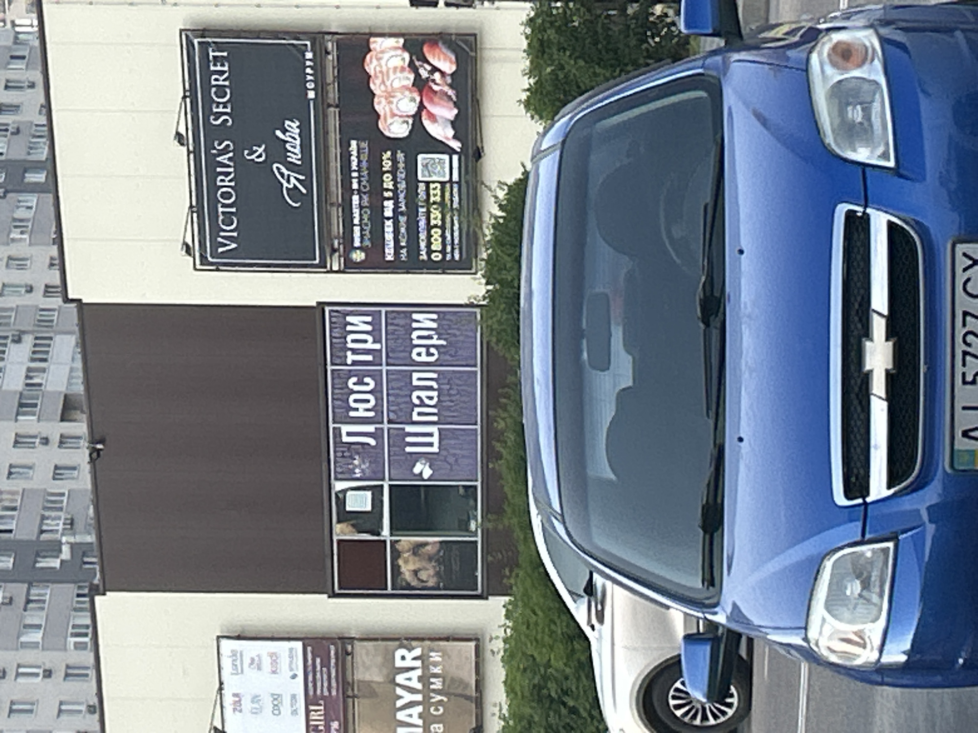 In Brovary, Ukraine, a blue Chevrolet Aveo is parked in front of a retail building with several signs. To the right of the Chevrolet, a silver SUV is parked. The retail building has a prominent sign for "Victoria's Secret". Below this sign is another sign advertising sushi with a phone number. To the left of the Victoria's Secret sign is a multi-pane window with some displays inside, and further left is a sign for "MAYAR" and "GIRL." The building is situated in a commercial area, with residential buildings visible in the background.