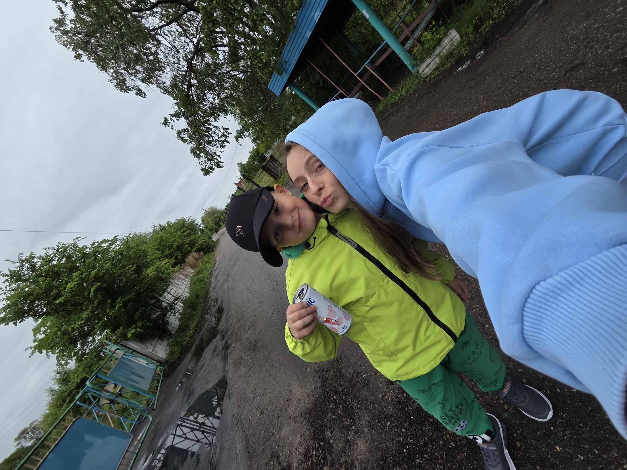 The image depicts two individuals on an unpaved road in Pronkino, Russia, under an overcast sky. A young boy is positioned on the left, wearing a black baseball cap with a white 'N' logo, a lime green waterproof jacket, dark green sweatpants with white graphics, and grey sneakers. He is holding a white aluminum can with a pink and red graphic, labeled "Lepic 0,25L," and is smiling directly at the camera. To the boy's right and slightly in front, a young woman or older teenager is present, wearing a light blue hooded sweatshirt with the hood up. She is looking at the camera, making a facial expression characterized by pursed lips. The ground consists of dark, unpaved earth with multiple puddles reflecting a blue metal gate or fence visible on the left side of the frame. Green foliage and trees line both sides of the path. In the background on the right, a small wooden structure with a blue roof and teal support beams, resembling a gazebo or covered bench, is visible.