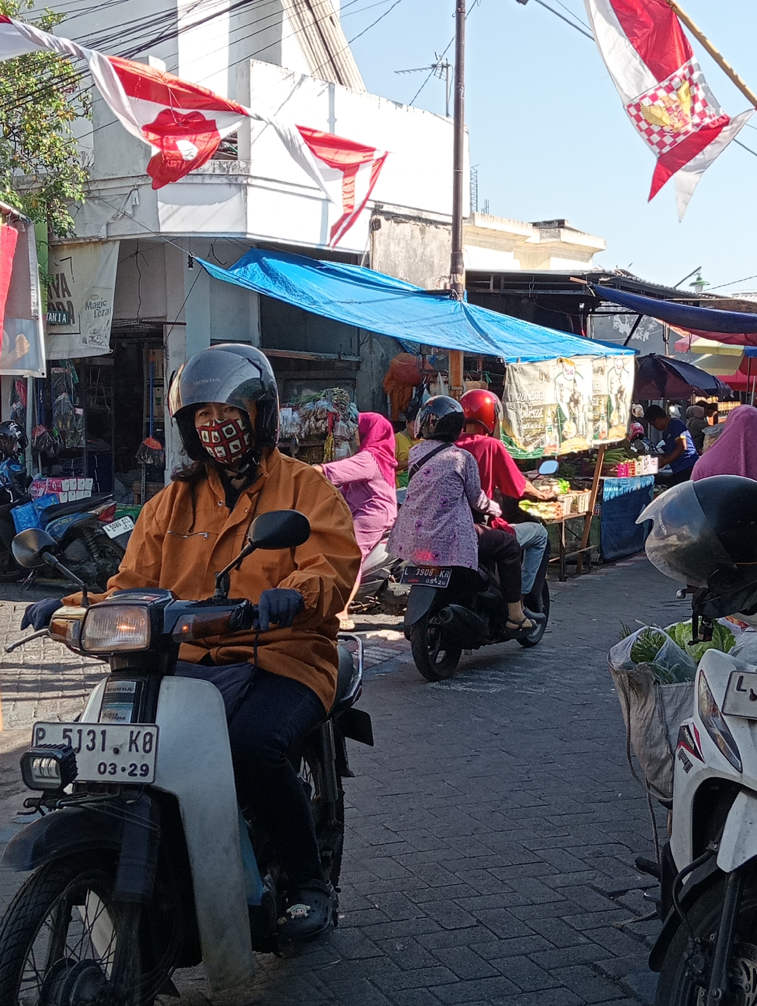 This is a daytime, eye-level outdoor photograph taken in Surabaya, Indonesia, likely in a market or street area, indicated by the presence of market stalls, awnings, and pedestrians on motorcycles.
In the foreground, a person wearing a helmet and a patterned face mask is riding a white motorcycle. They are wearing an orange jacket and dark pants, with dark gloves on their hands. The motorcycle has a license plate that reads "P 5131 KO 03-29".
Behind the first motorcycle, another motorcycle carries two passengers. The person in the back is wearing a red helmet and a red shirt, while the person in front is wearing a purple patterned top and a pink headscarf.
Further in the background, various market stalls are visible, covered by blue and dark tarps. People can be seen browsing or working at the stalls. Several other motorcycles are parked or being ridden in the scene. Red and white flags, likely Indonesian flags, are strung across the top of the frame, suggesting a national celebration or event. The ground is paved with interlocking bricks. The overall impression is a busy, everyday scene in a bustling urban environment.