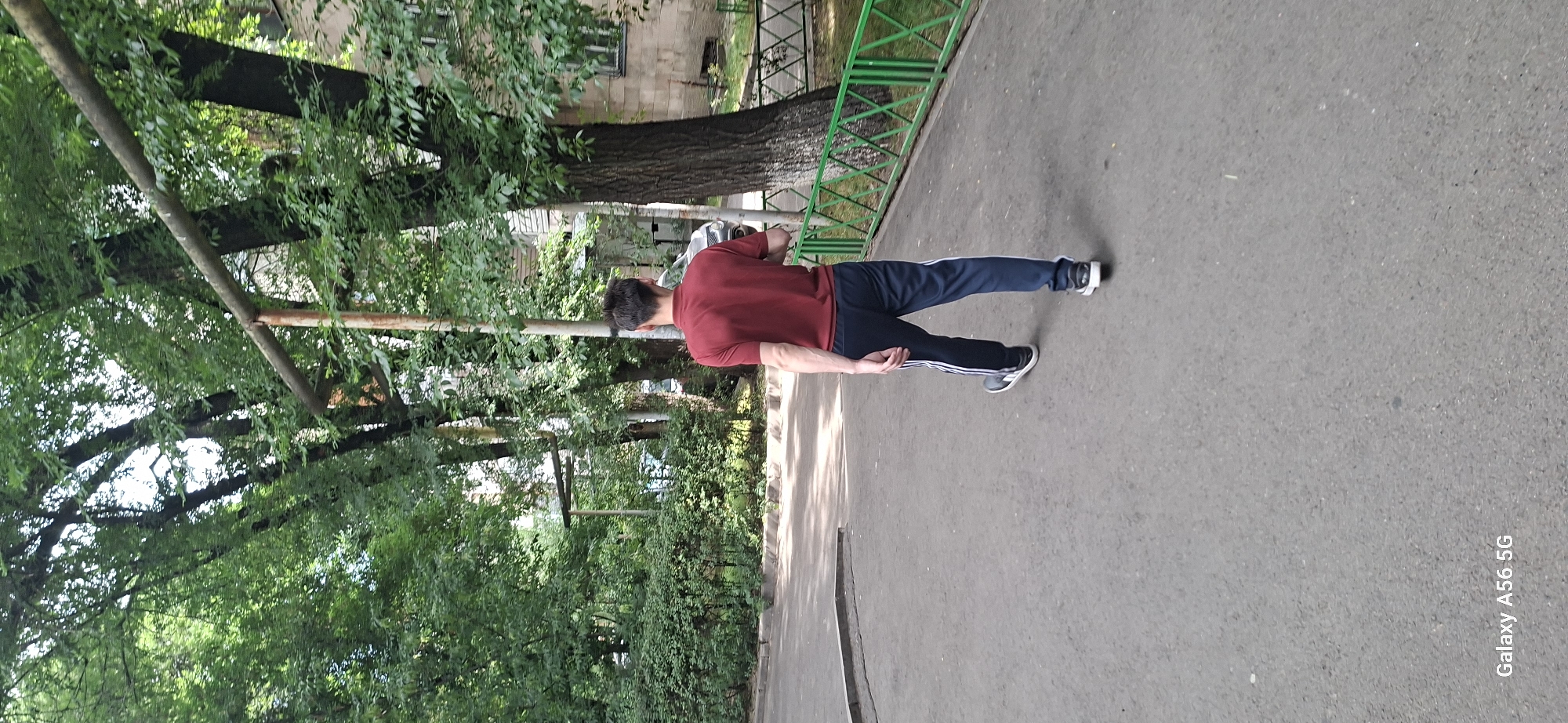 A man in a red polo shirt and dark blue athletic pants with white stripes walks away from the camera on a paved path. He is wearing black and white sneakers. The background features lush green trees, a building with windows, and a green metal fence. The scene appears to be an outdoor area in a residential or park-like setting, potentially in Almaty, Kazakhstan, given the "Galaxy A56 5G" watermark, which is a model of Samsung phone often used by residents in the region. The lighting suggests it is daytime.
