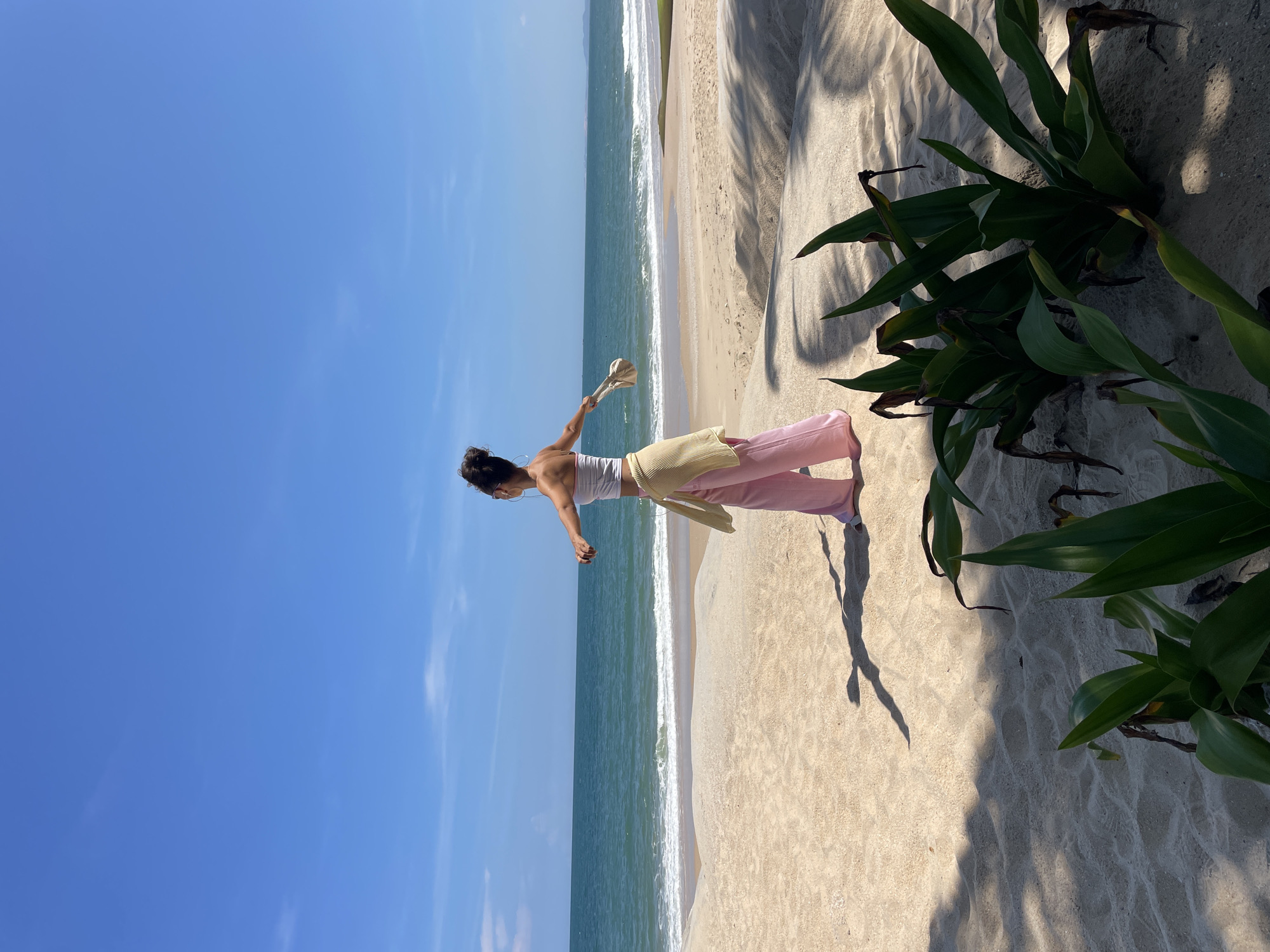 A person stands on a sandy beach, facing the ocean. The person is wearing a white crop top, pink pants, and a yellow cardigan tied around their waist. They are holding a light-colored bag in their right hand and have their arms outstretched to the sides. The ocean is calm with small waves, and the sky is a clear blue with a few wispy clouds. Green plants are visible in the foreground, casting shadows on the sand. The overall scene suggests a relaxed day at the beach in Phu Thuy Ward, Vietnam.