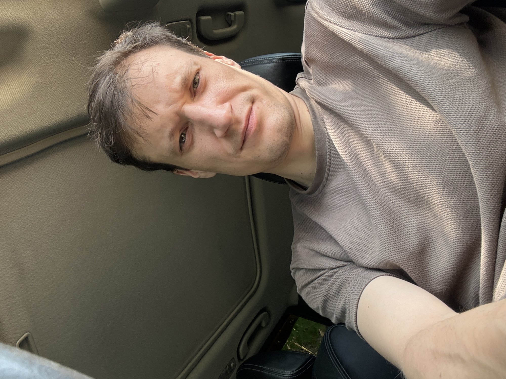 A man is taking a selfie inside a car. He is seated and his face is angled towards the camera. The man has light skin, brown hair, and green eyes. He is wearing a tan t-shirt. The interior of the car is visible, including a sunroof and leather seats. Sunlight is streaming into the car, casting shadows on the man's face and t-shirt. The background shows trees and foliage, suggesting the car is parked in a natural setting, possibly outside the city of Makiv, Ukraine.