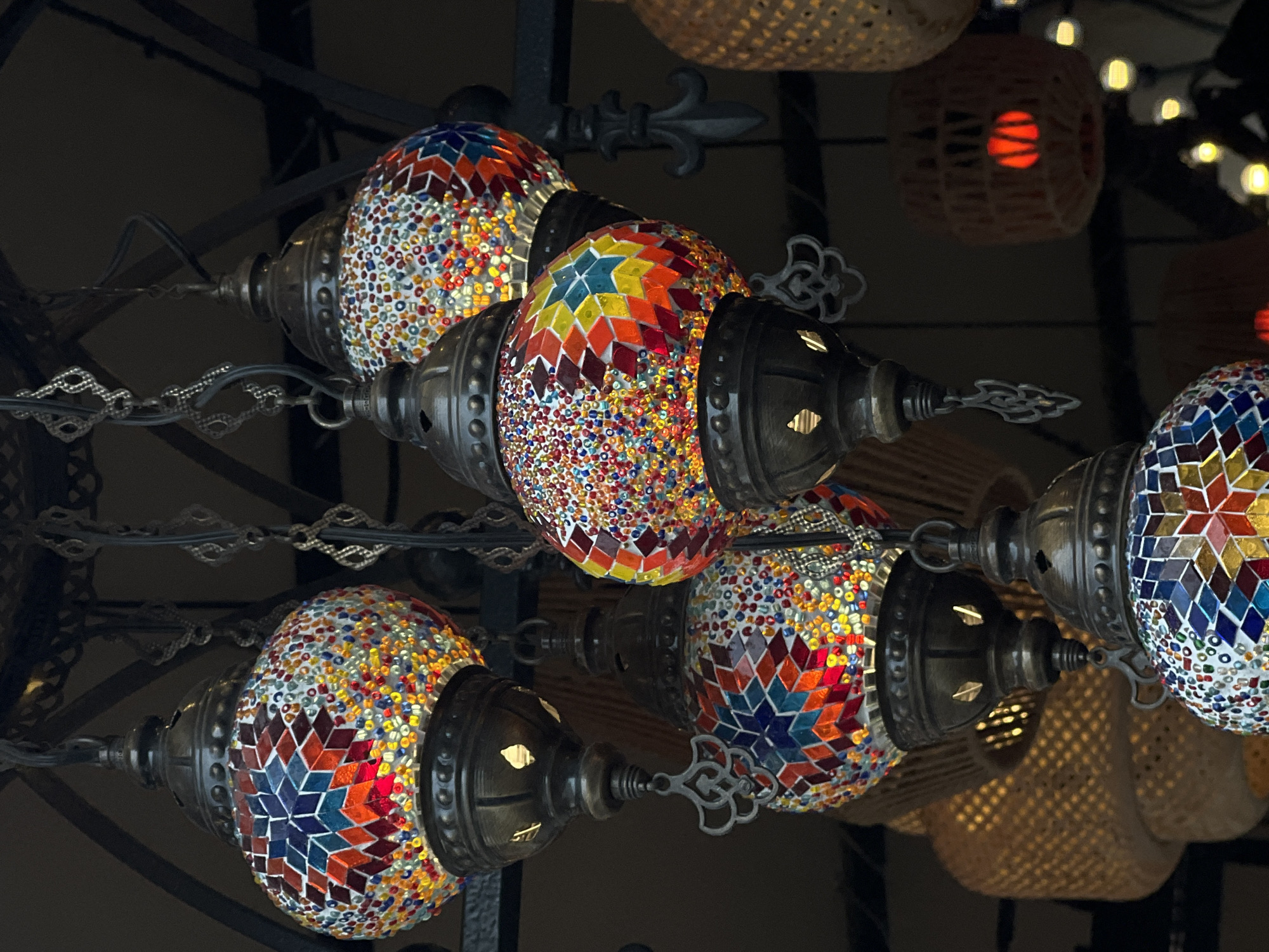 A detailed image shows a collection of ornate, multicolored Turkish mosaic lamps hanging from a wrought iron fixture. The lamps are egg-shaped, covered in small, brightly colored mosaic tiles arranged in geometric patterns. The metal casings of the lamps appear to be aged, with a dark, antiqued finish and intricate detailing. In the background, there are woven basket-like light fixtures, some emitting a soft, warm light. The overall scene is dimly lit, creating a sense of ambiance. No people or specific actions are visible in the image. The location is listed as the city of Unknown, Unknown.