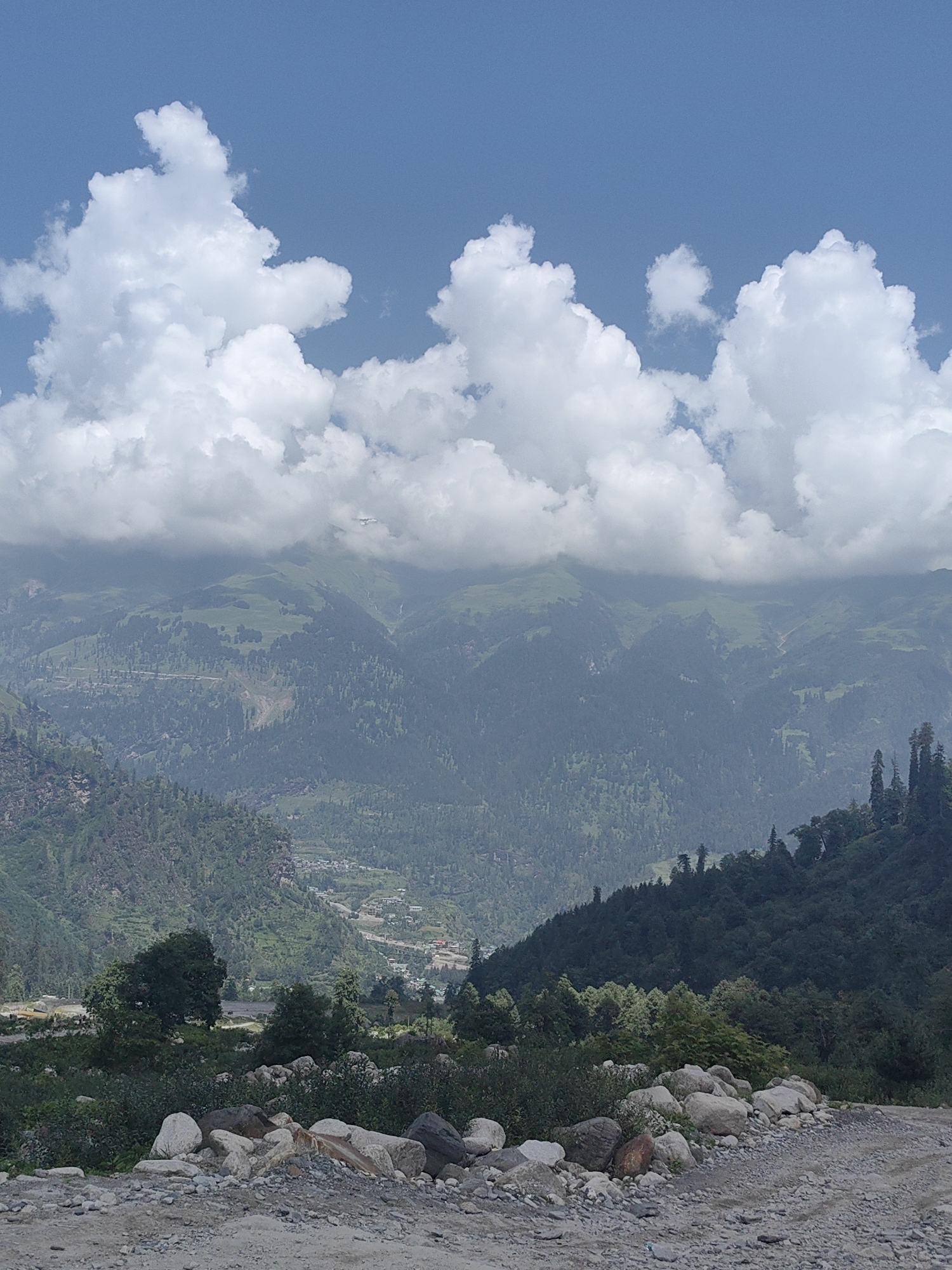 The image depicts a vast mountainous landscape under a bright blue sky with fluffy white clouds. The foreground features a rough, gravelly path strewn with large, grey, and white rocks, some smooth and others angular. Lush green vegetation, including bushes and trees, flanks the path, with larger trees visible further into the scene.
In the mid-ground, rolling green hills are densely covered with a mix of dark green coniferous trees and lighter green foliage. A winding road is visible traversing one of the hillsides. A small village or settlement with buildings can be seen nestled in a valley between the mountains. The background consists of further mountain ranges, with the highest peaks appearing hazy and distant. A faint waterfall can be spotted on one of the distant slopes. The overall impression is one of natural beauty and a rural, mountainous setting in Majhach, India. No people are visible in the image.